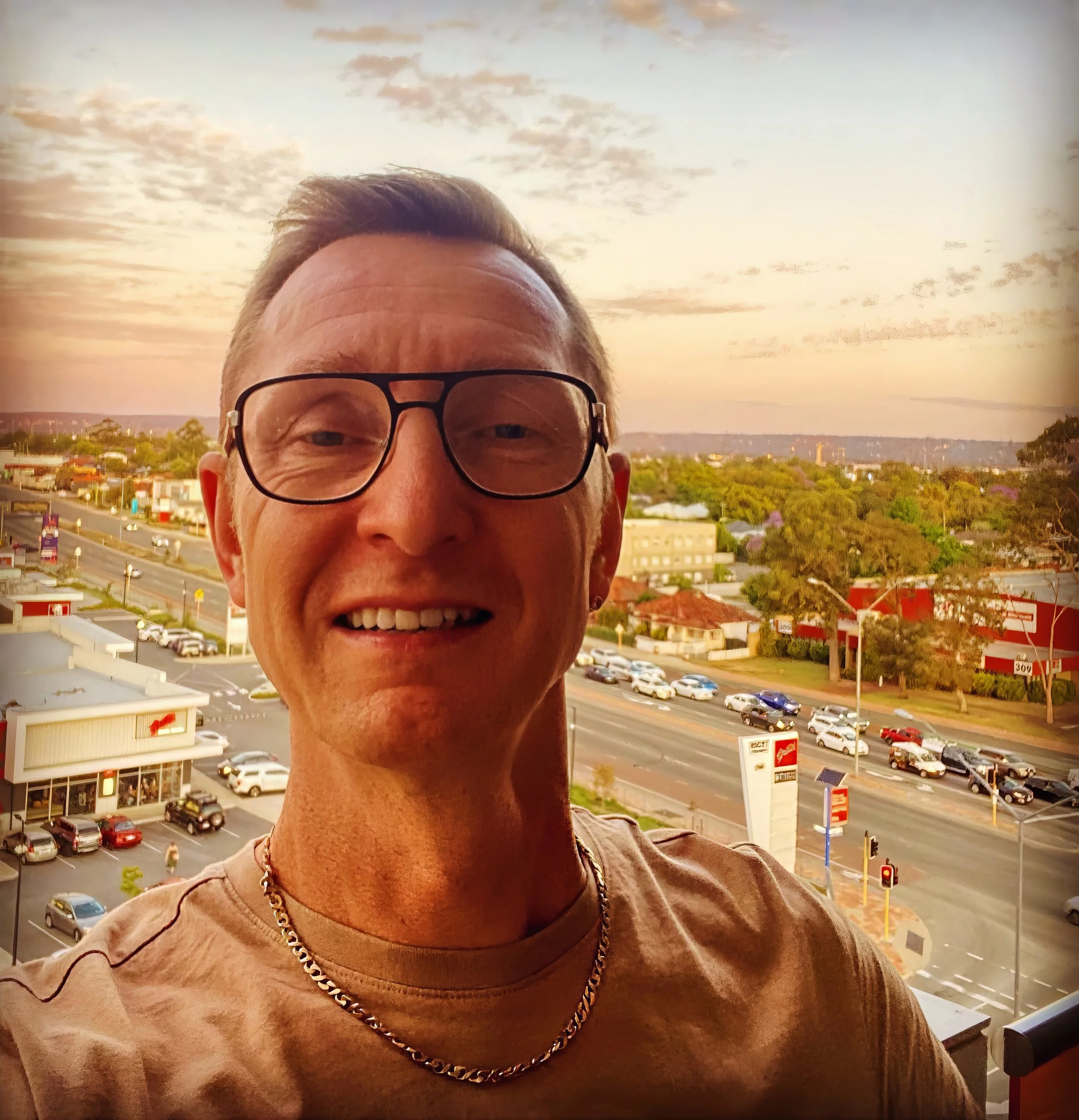Dean Charles wearing glasses and a chain necklace takes a selfie on a balcony overlooking a busy street at sunset with a clear sky and scattered clouds.