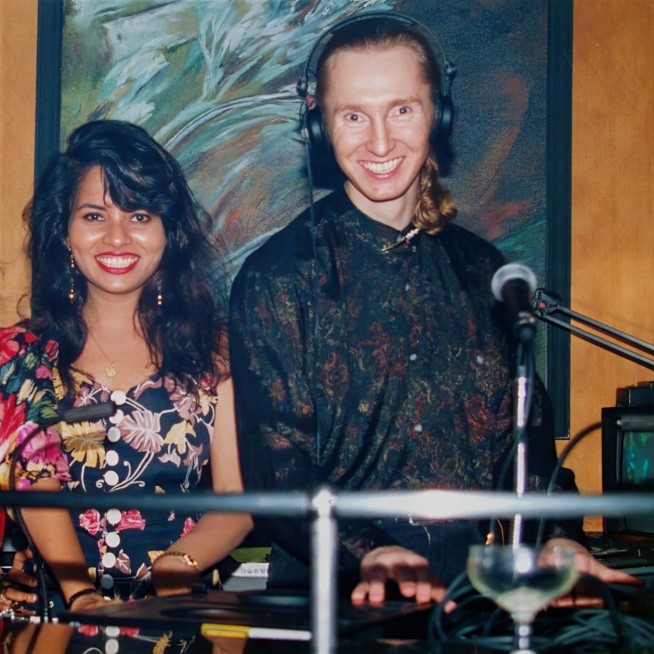 Two people smiling and posing for a photo. The man on the right is wearing headphones and is standing behind a microphone, with a turntable DJ equipment in front of him. The woman is wearing a colorful floral dress and jewelry. There is a painting in