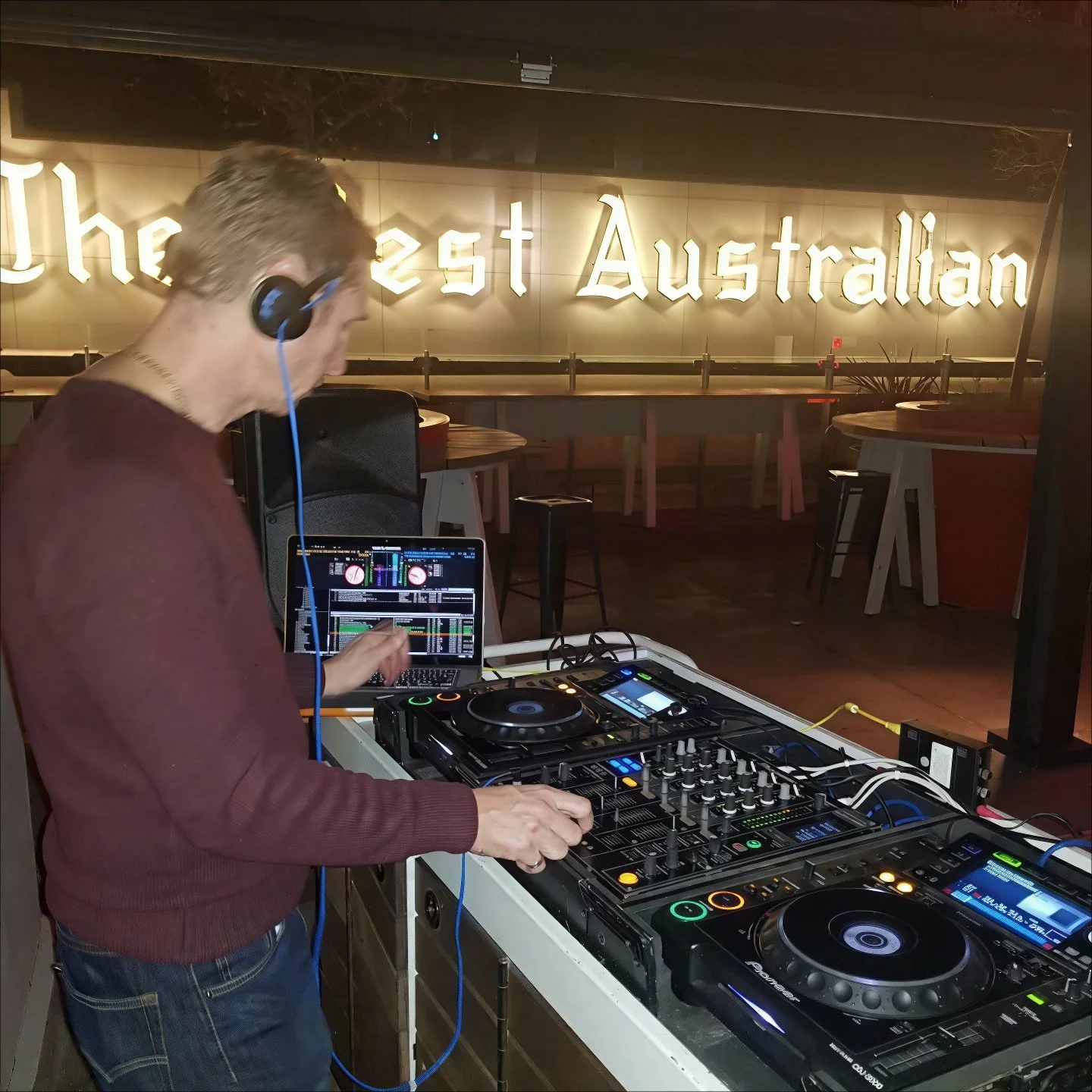 DJ Dean Charles in a maroon sweater and jeans mixing music on professional DJ equipment with a laptop, in front of a large illuminated sign reading 'The West Australian' at Print Hall bar in Perth, Western Australia.