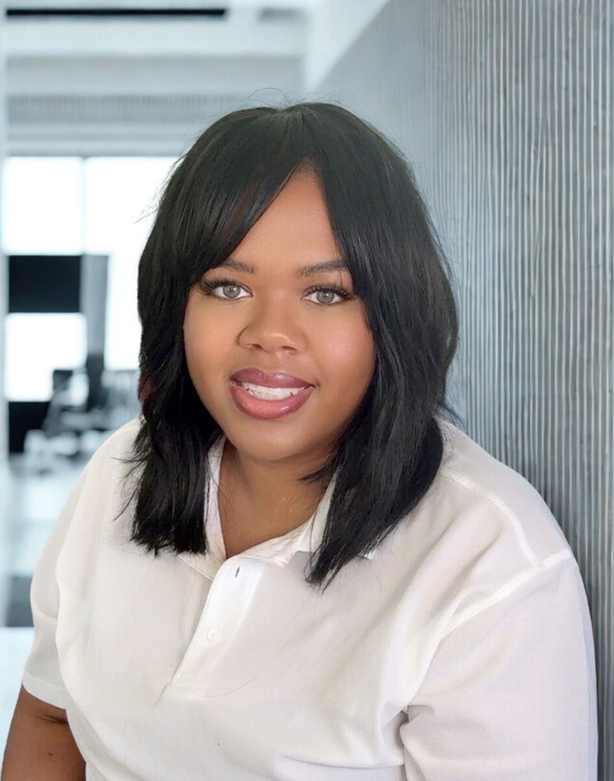 A woman with black hair and light skin smiling, wearing a white shirt, sitting indoors.