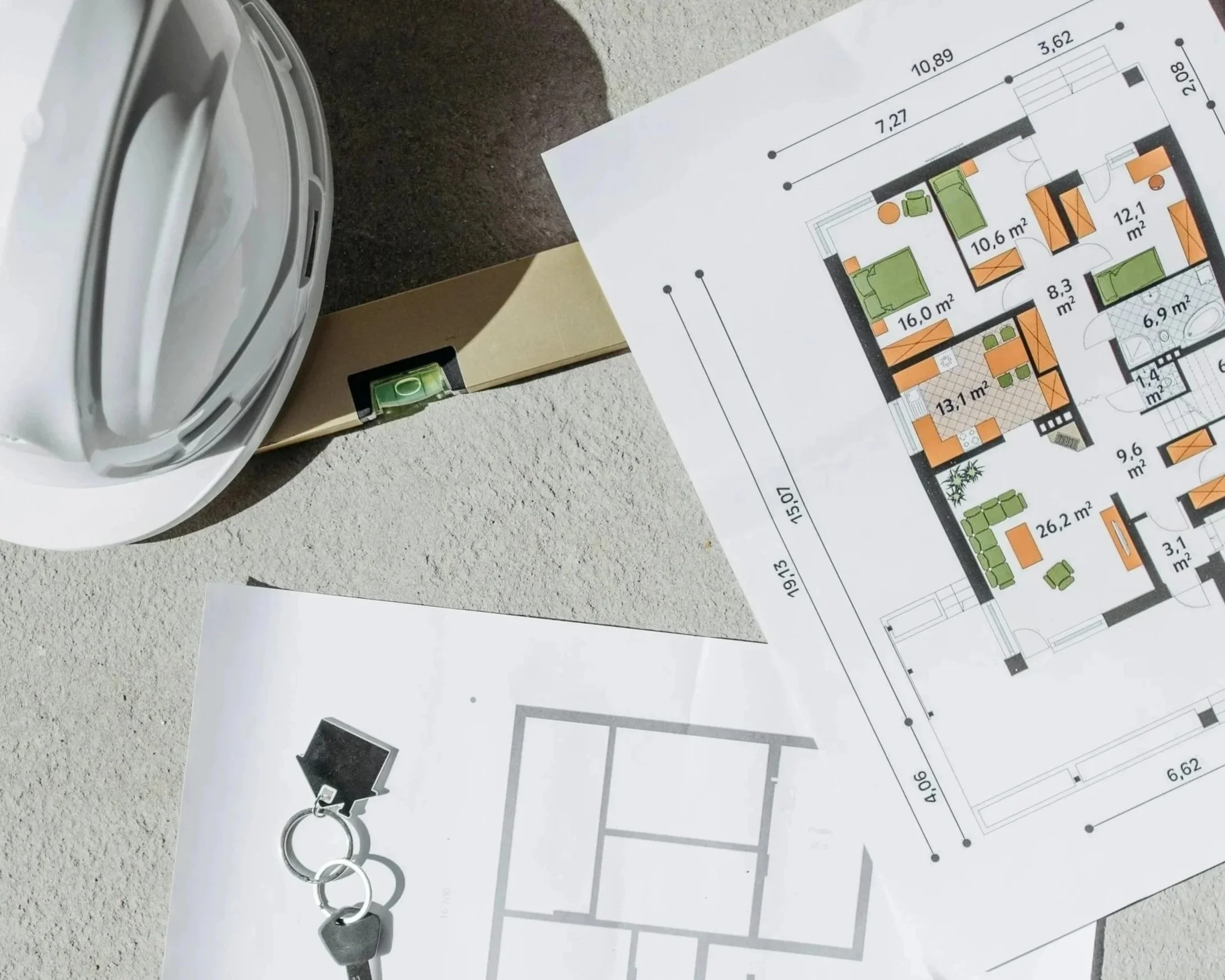 Architectural blueprint of a house floor plan, a white hard hat, a key with a keychain, and a spirit level on a construction site.