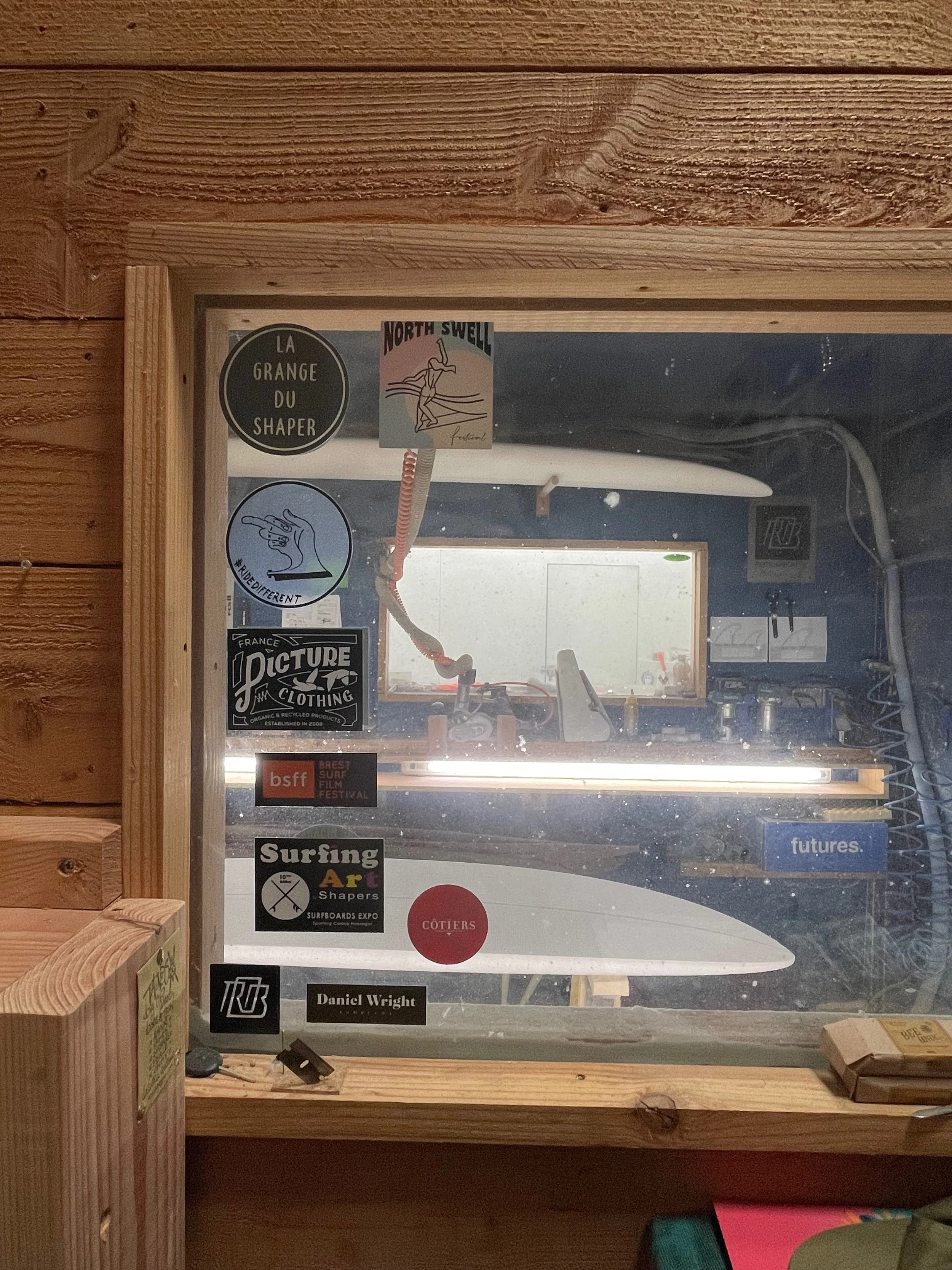 Vitrine en bois avec plusieurs autocollants liés au surf, dont 'La Gange du Shaper', 'North Swell' festival, 'Picture Organic Clothing', 'Brest Surf Film Festival', 'Surfing Art by Shapers', 'Swing Surf'