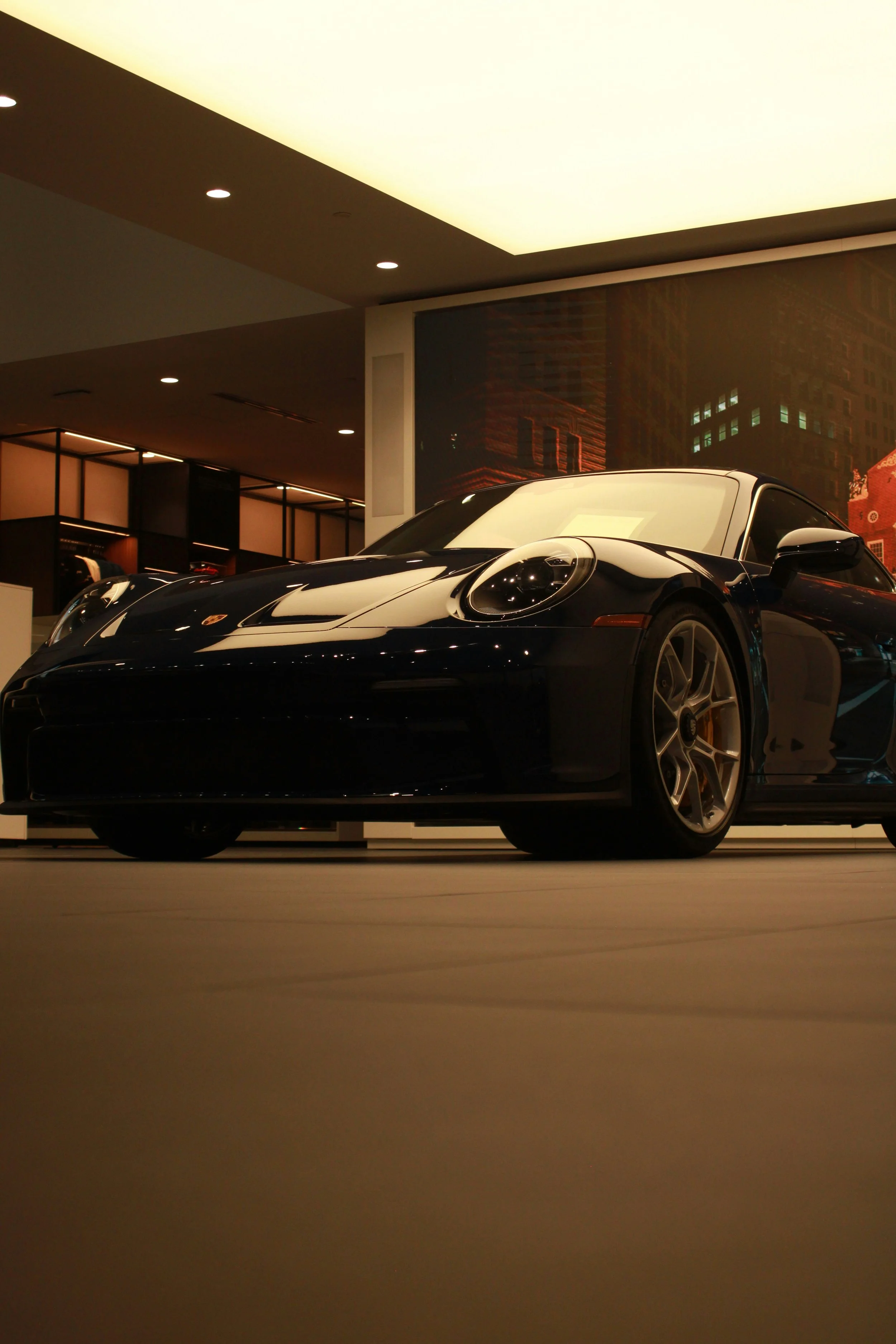 A black sports car on display indoors, with bright lights overhead and a cityscape mural in the background.