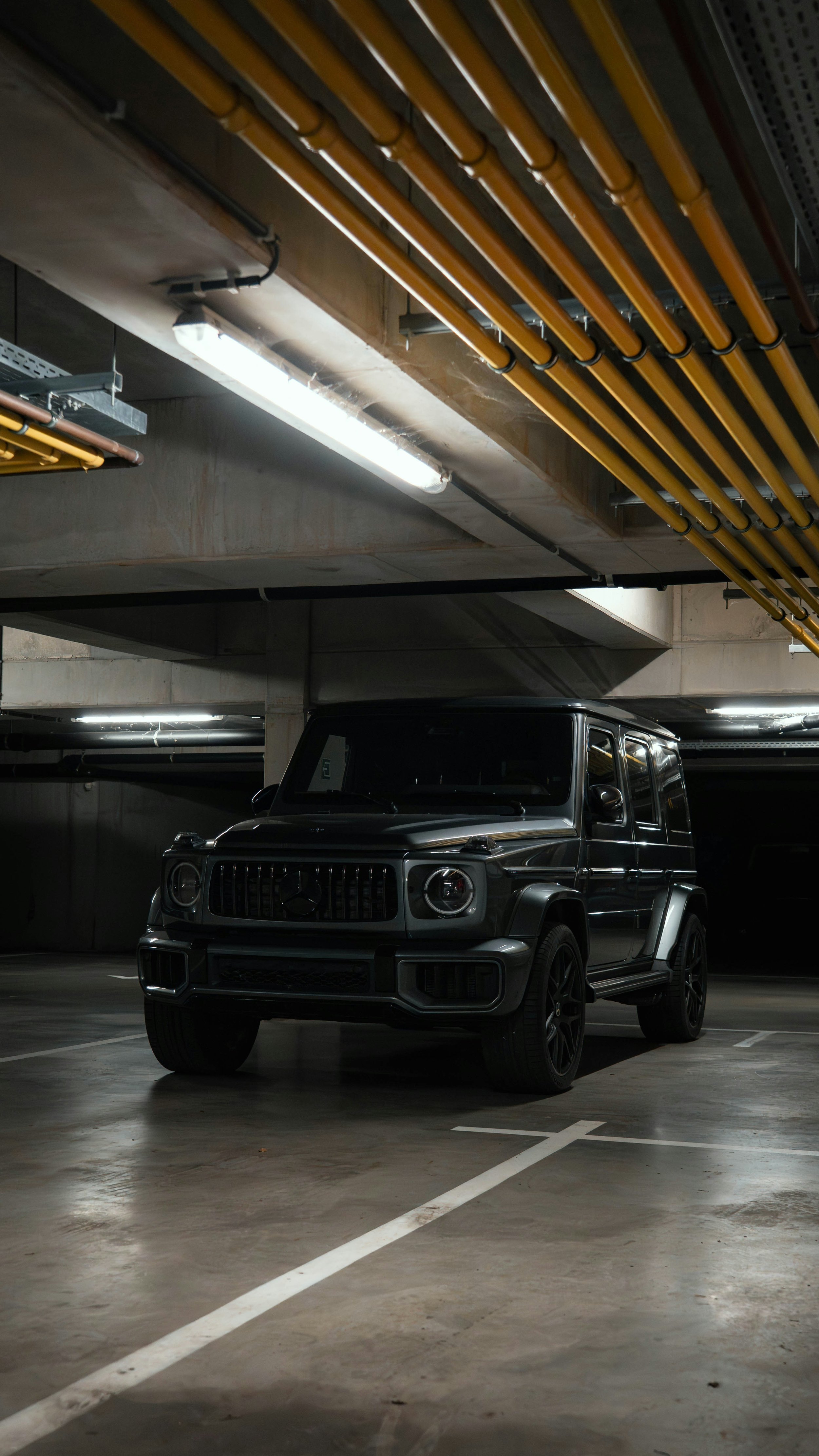 A black Mercedes-Benz G-Class SUV parked in an underground parking garage with overhead lighting and exposed pipes.