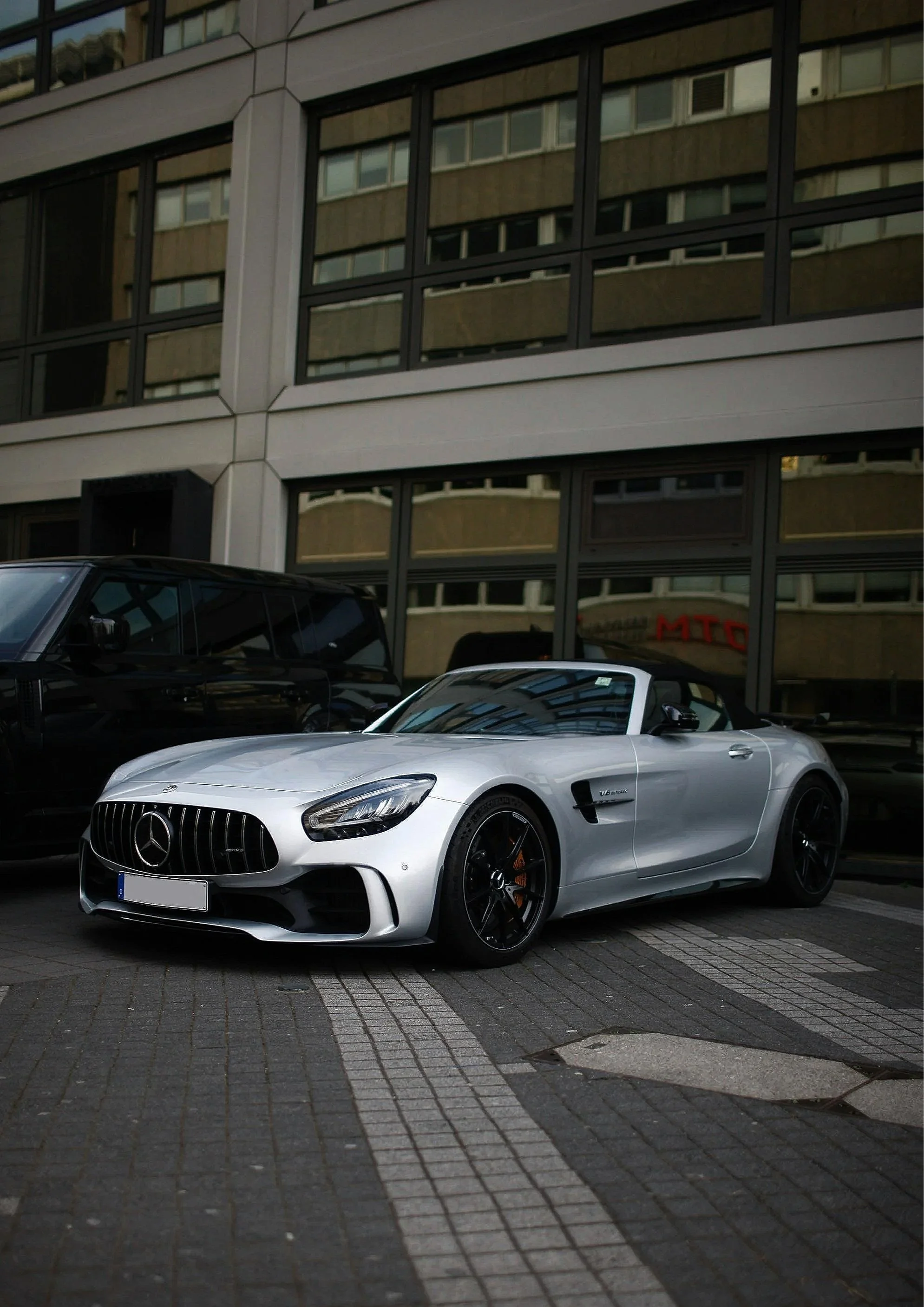 A silver Mercedes-Benz AMG GT sports car parked on a street in front of a modern glass building.
