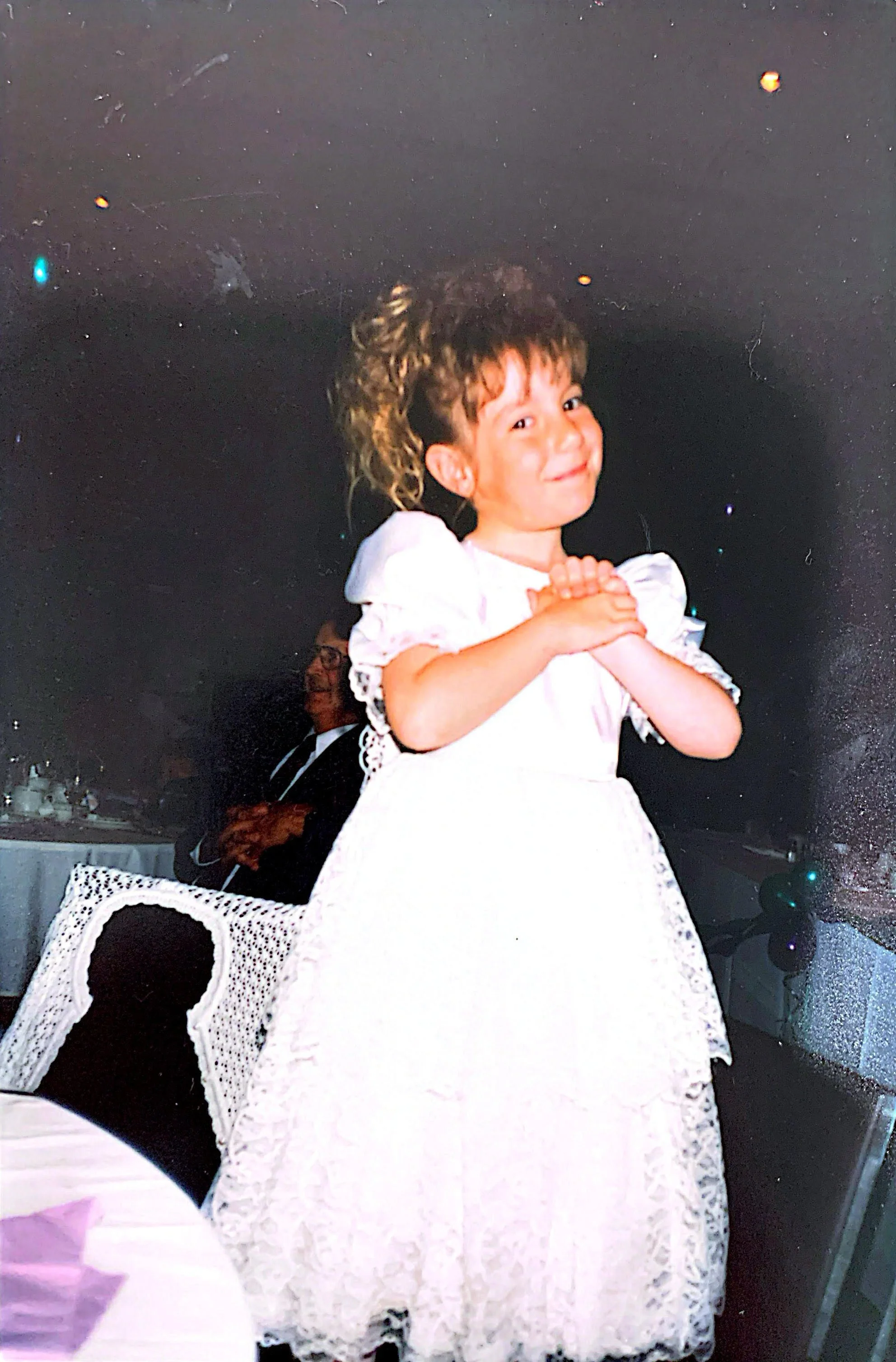 Une jeune fille souriant avec les mains jointes, portant une robe blanche en dentelle, dans un cadre festif, avec d'autres personnes et décorations en arrière-plan.