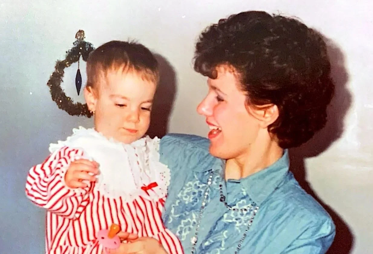 Une femme tient un bébé fille dans ses bras. La jeune fille porte une robe à rayures rouges et blanches avec une décoration en dentelle et un petit ruban rouge. La femme porte une chemise en denim et a des cheveux bouclés foncés.