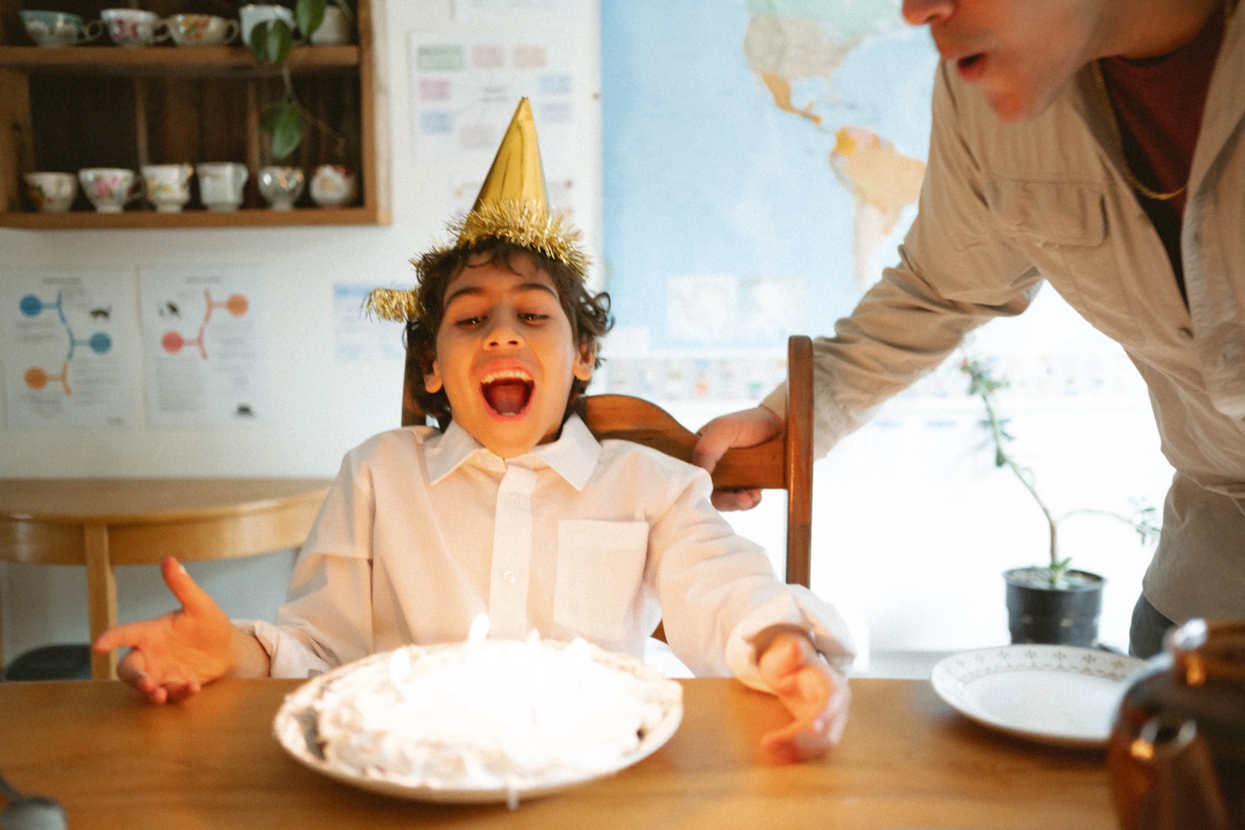 Un garçon avec un chapeau de fête sourit avec la bouche ouverte à une fête d'anniversaire. Il est assis à une table avec un gâteau d'anniversaire et des assiettes. Un adulte est à côté de lui, peut-être en train de lui faire une surprise.