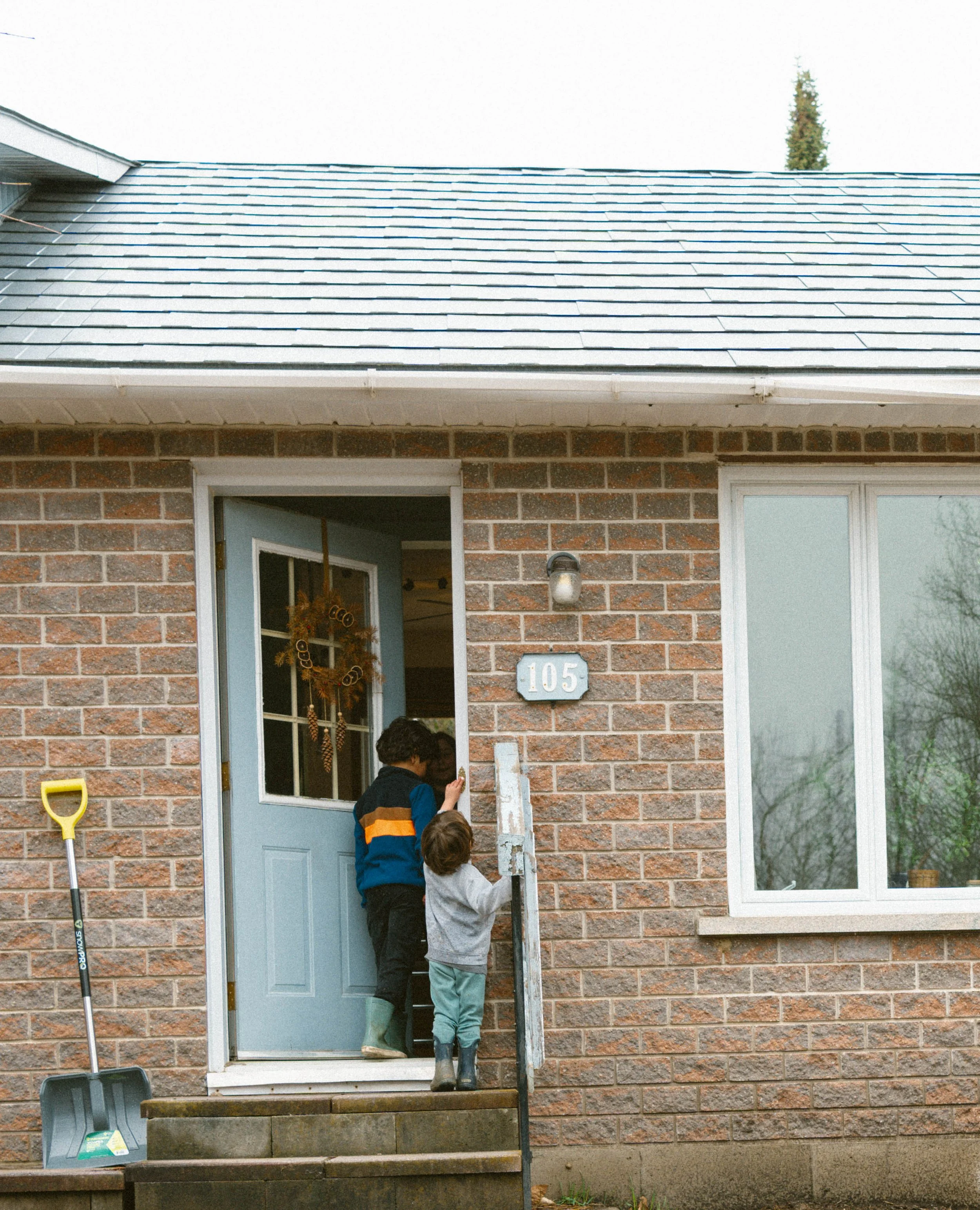 Deux garçons sonnent à la porte d'entrée d'une maison en briques, discutant et souriant, avec des outils de jardinage à côté.