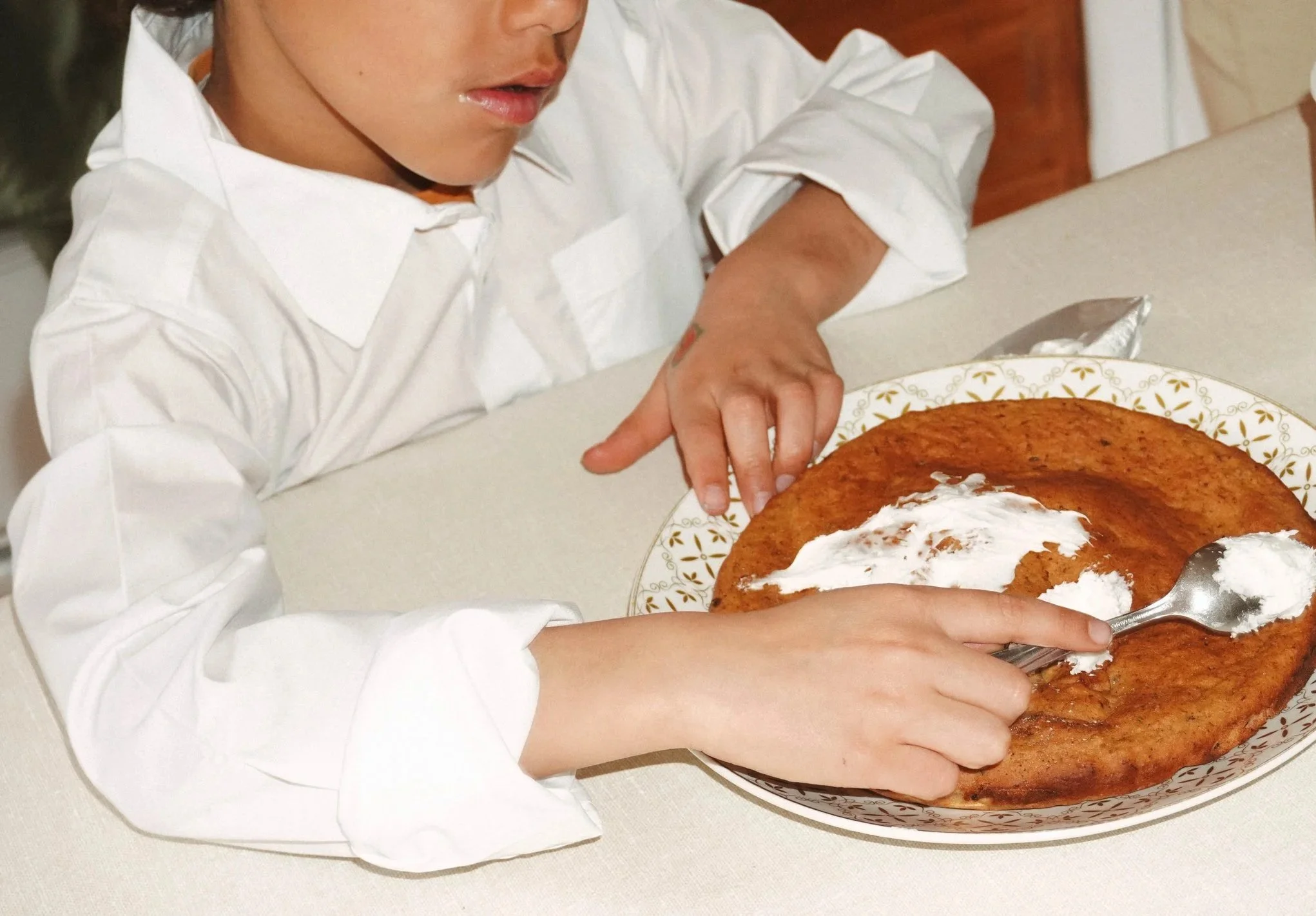 Une personne crème un gâteau d'un crèmage à la vanille dans une assiette décorée, portant une chemise blanche.