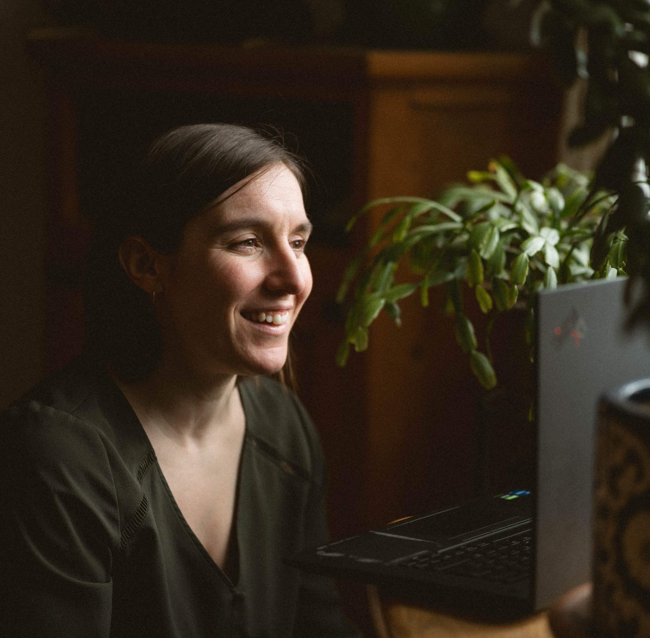 Une personne avec des cheveux bruns, souriante, regarde un écran d'ordinateur dans un environnement intérieur faiblement éclairé.