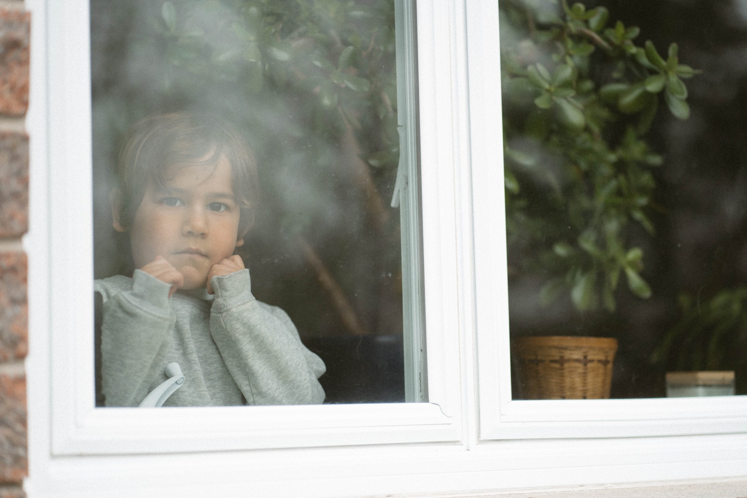 Un enfant regarde par une fenêtre à double vitrage, avec une expression pensive. Il porte un sweat gris clair et se tient près de la fenêtre, à l'intérieur d'une maison.