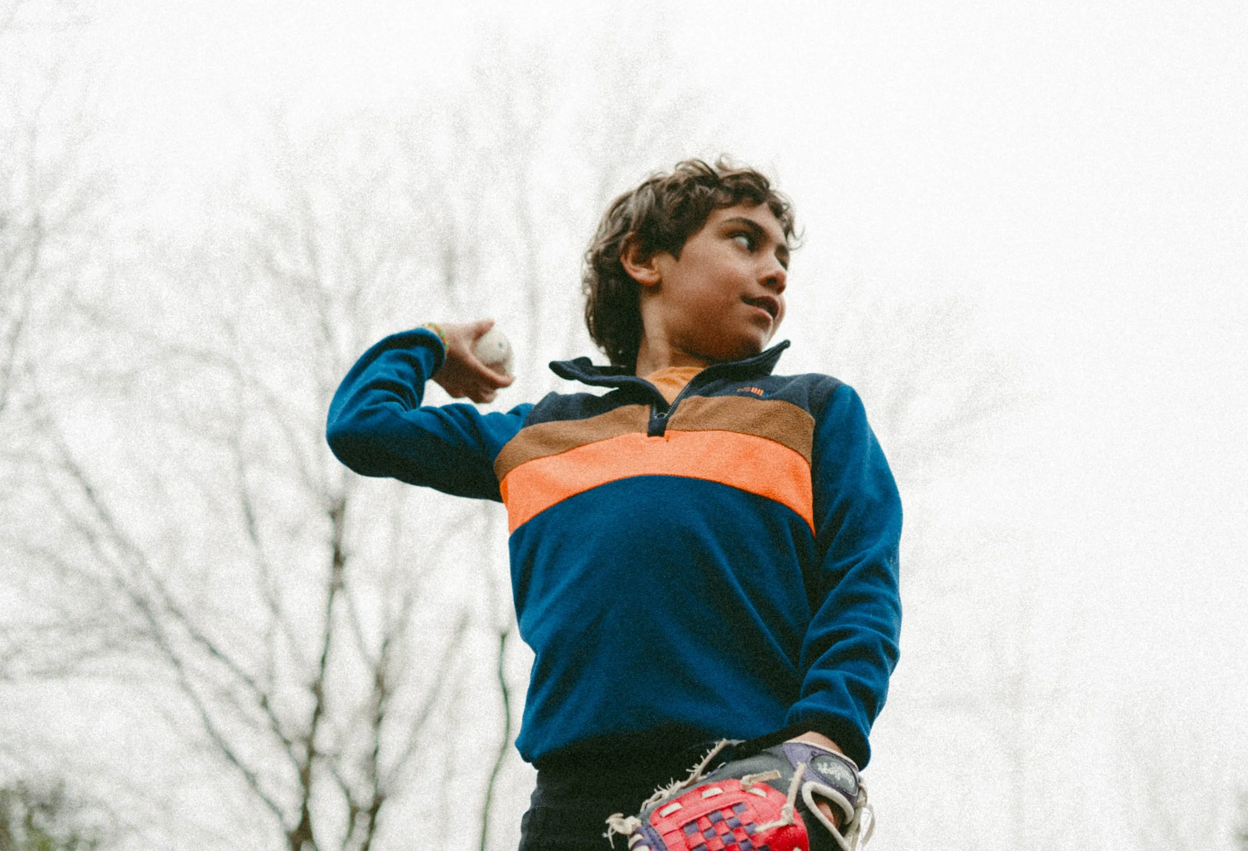 Un jeune garçon en train de jouer au baseball, portant une veste colorée et une chaussure de sport, en plein air avec un ciel nuageux et des arbres en arrière-plan.
