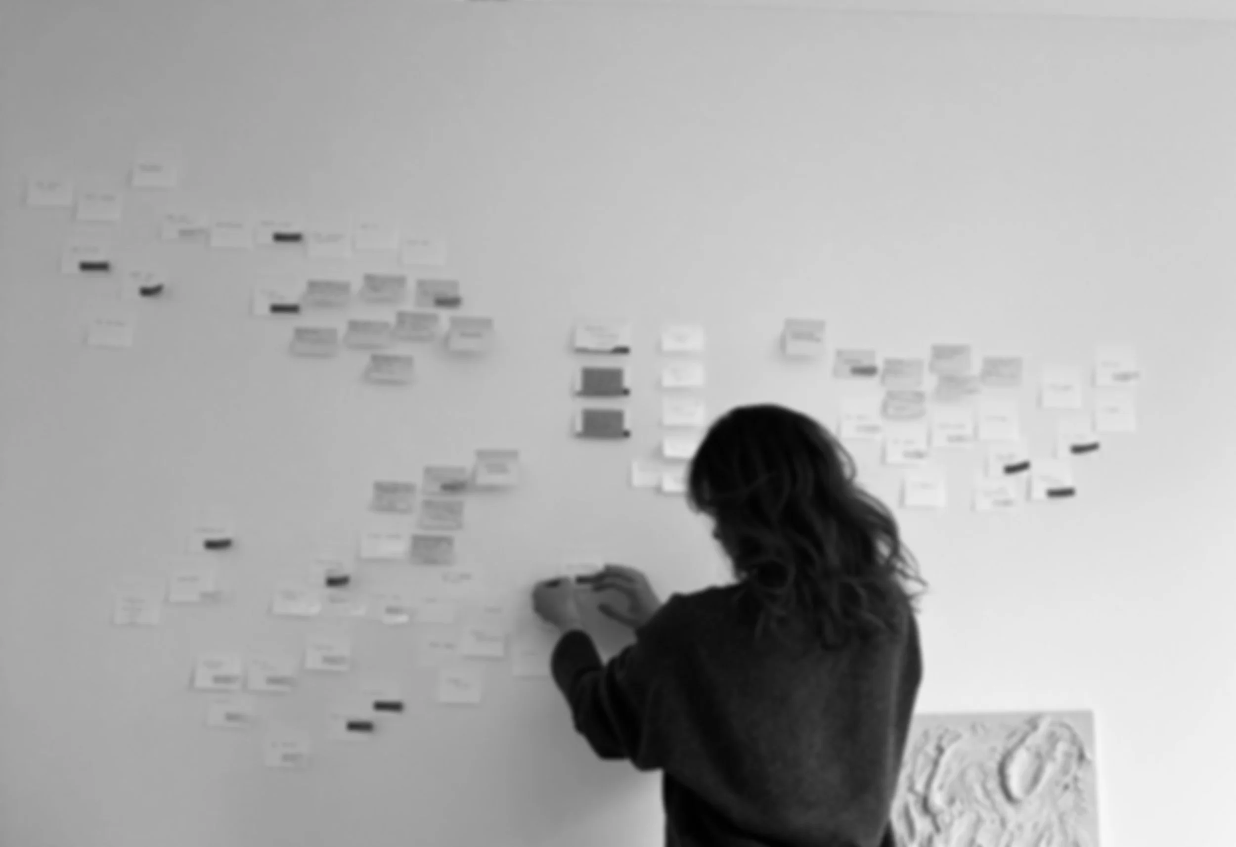 Woman with wavy hair standing in front of a wall with numerous sticky notes attached, working or organizing.
