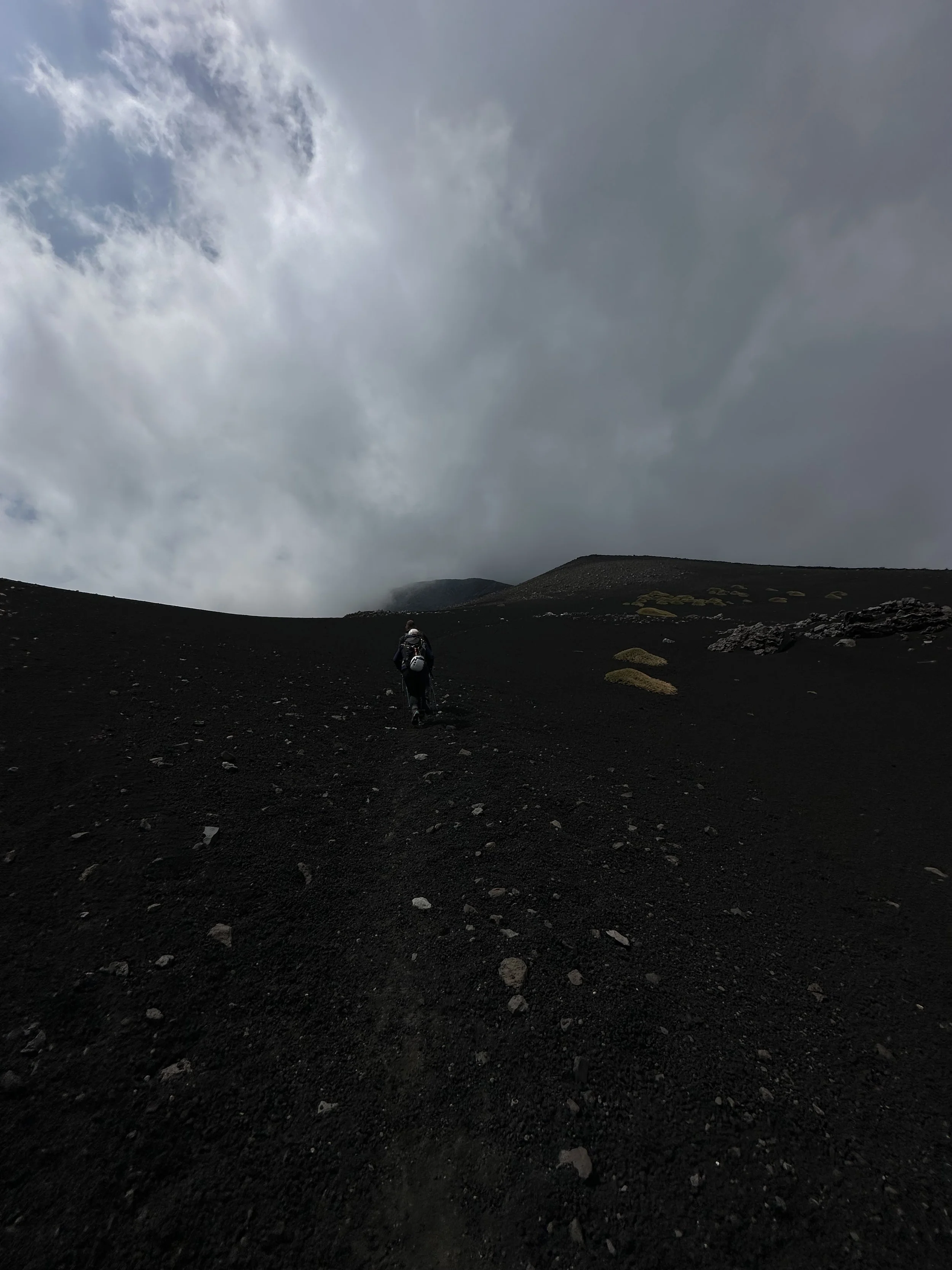 A person hiking up a dark, rocky slope towards a cloudy sky, with some sparse green vegetation on the ground.