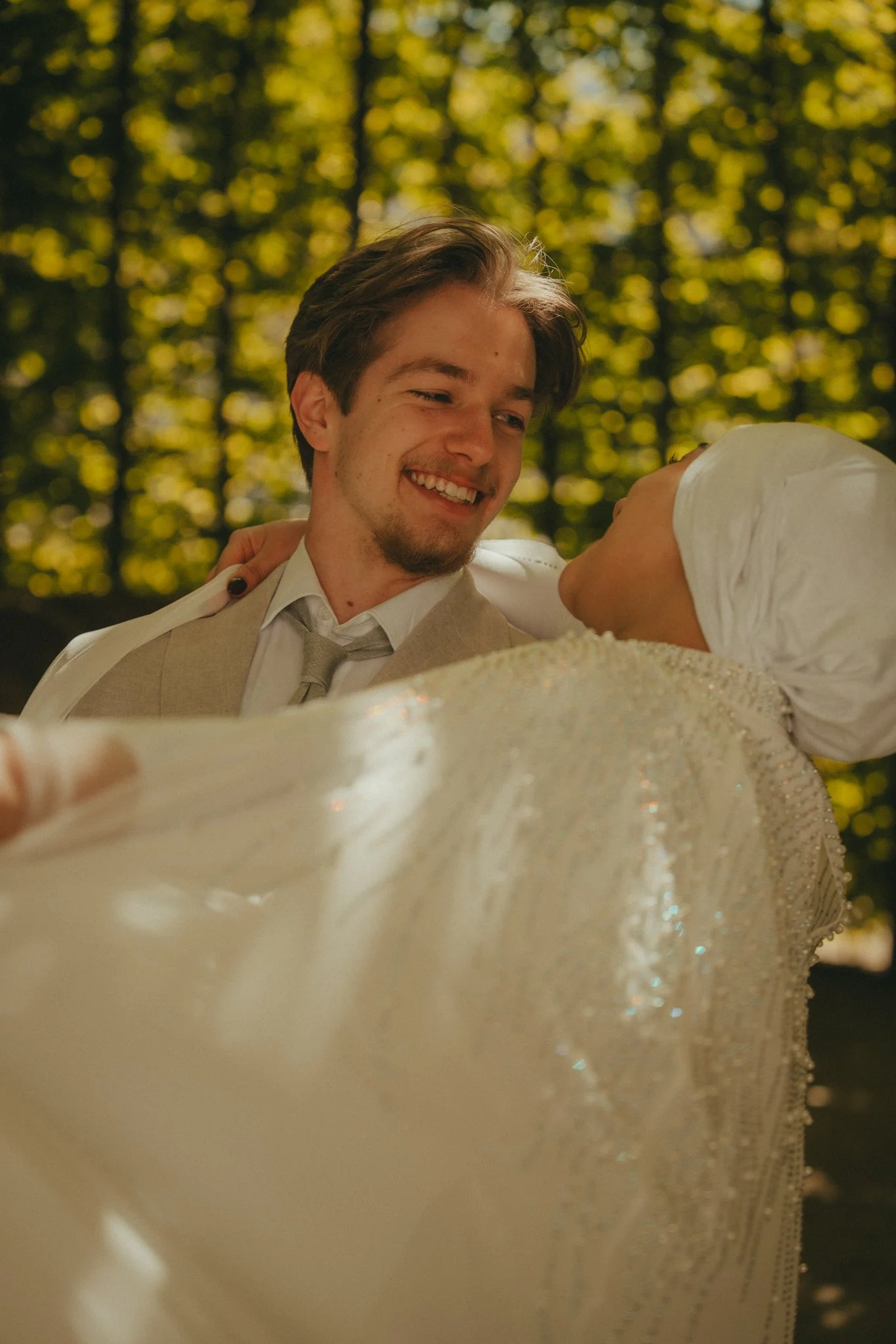 A smiling young man in formal attire, holding a woman in a white dress, in an outdoor setting with trees and yellowish-green foliage in the background.