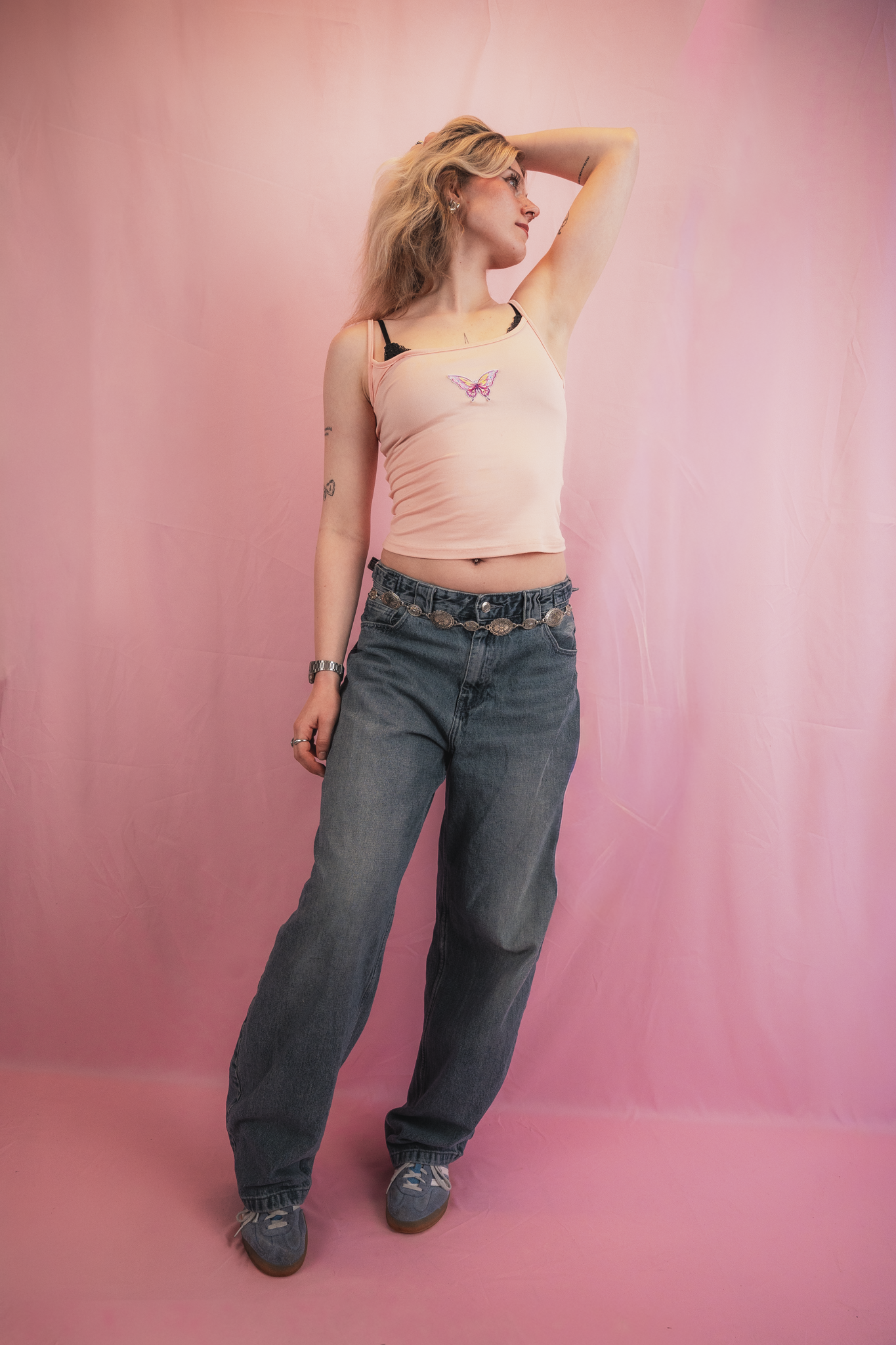 A woman with blonde hair standing against a pink backdrop, wearing a pink tank top with an embroidered butterfly, blue jeans, and sneakers, with her left arm raised and her head turned slightly to the left.