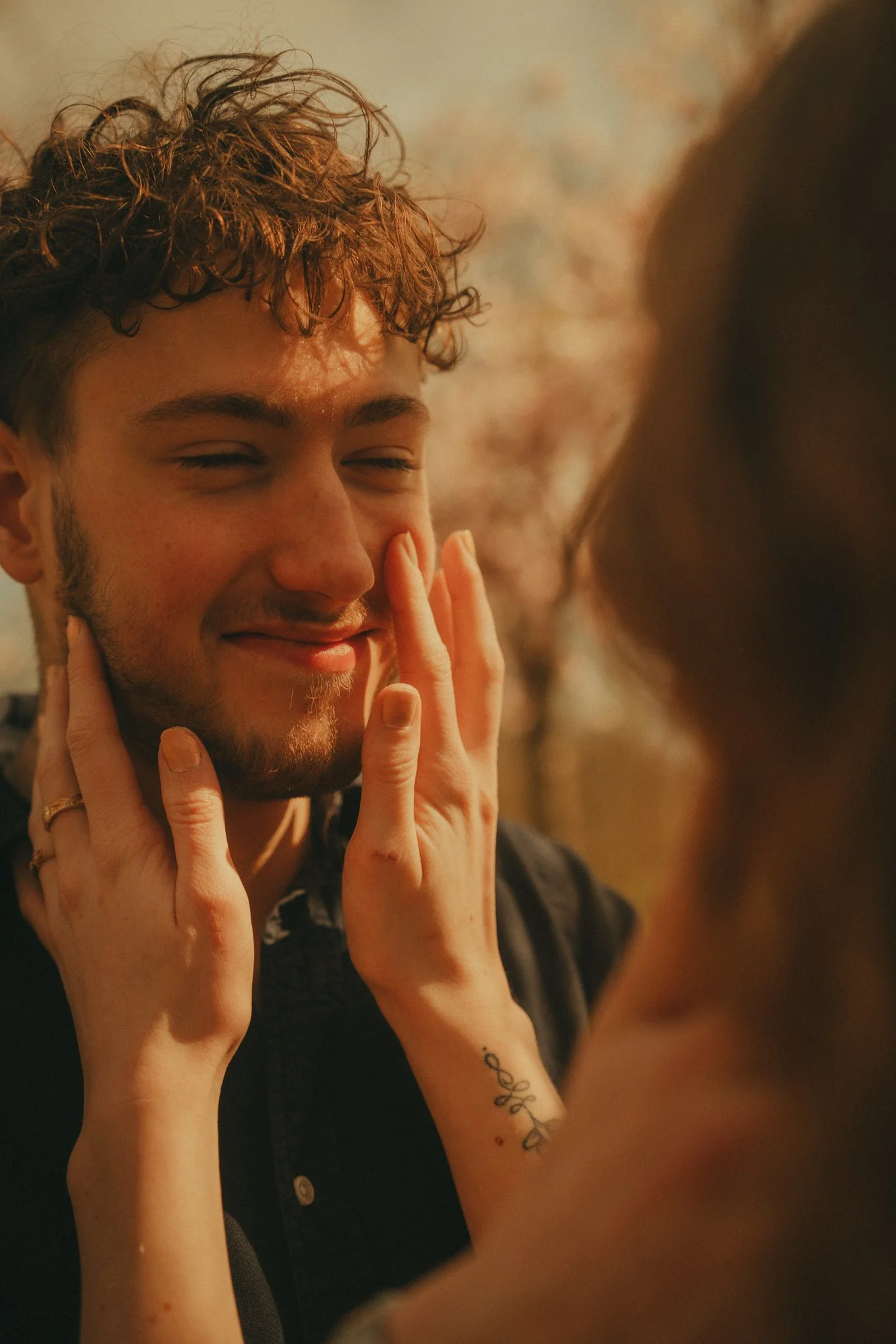 A young man with curly brown hair and a beard is smiling with his eyes closed. Someone touches his face gently, with fingers on his cheek and chin. The background is blurred with warm tones.