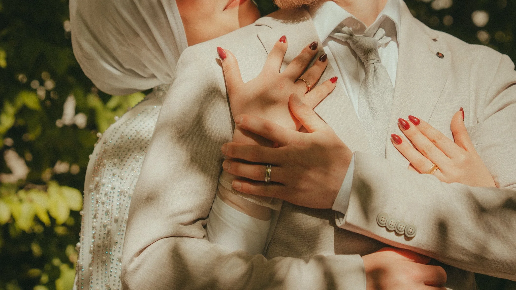 A close-up of a couple embracing, with the woman’s hands on the man's chest. They are dressed in light-colored formal attire, with the woman's nails painted red and gold. The background is blurred greenery.