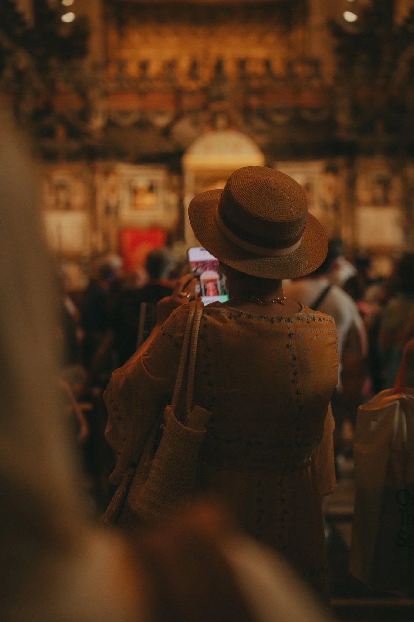 A woman wearing a wide-brimmed straw hat and a yellow patterned shirt, holding a phone and taking a photo in a crowded indoor venue with traditional, ornate architectural decor.