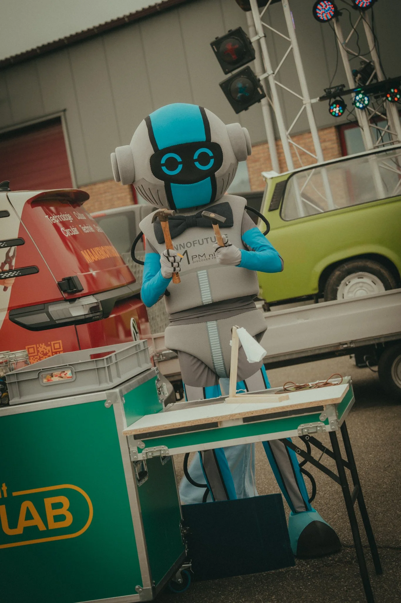 A robot with a round head resembling a gaming controller, wearing a gray suit and black bow tie, holding a hammer, standing at an outdoor event with trucks, tables, and stage lighting in the background.