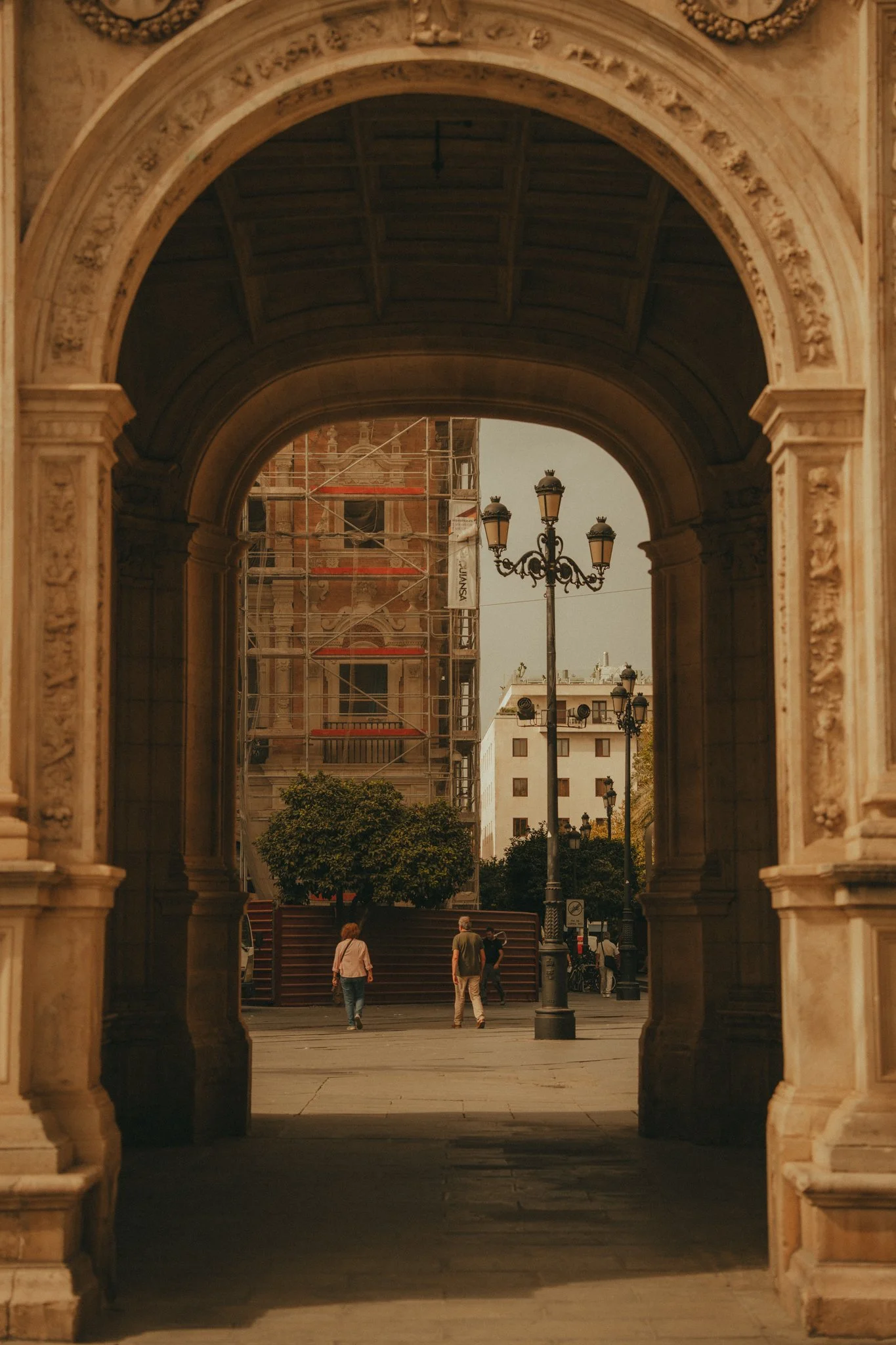 People walking through a city archway with street lamps and trees in the background, buildings around, and a building under renovation with scaffolding.