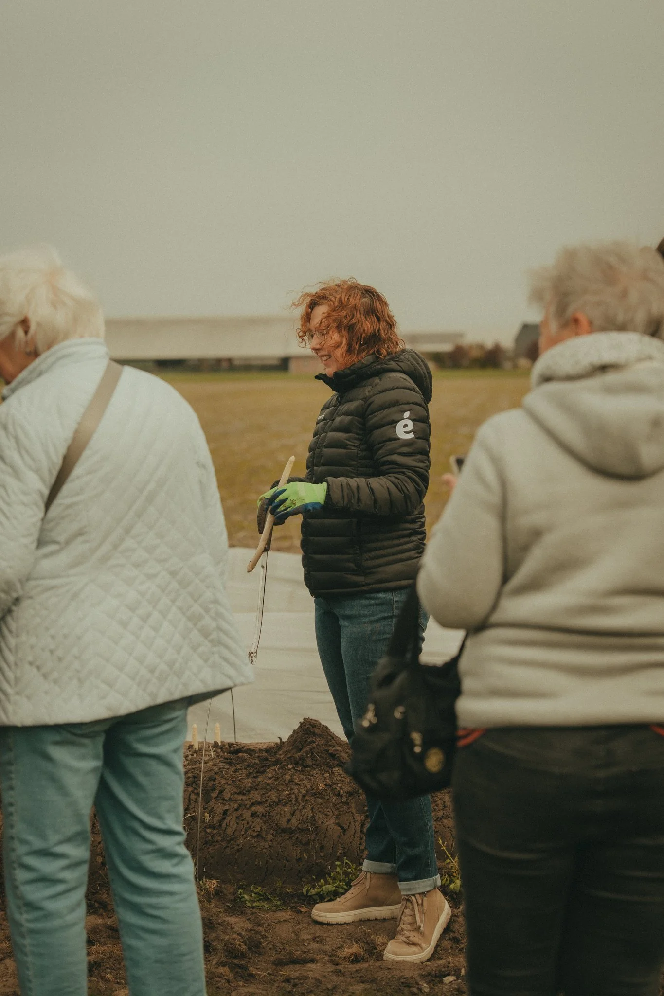 Group of people standing outdoors on a cloudy day, some with gloves, near a large mound of dirt.