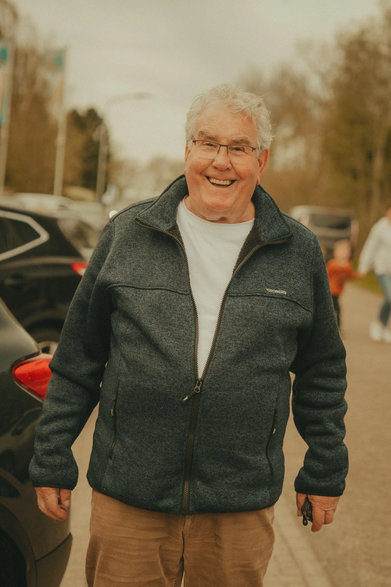 A smiling elderly man with gray curly hair, glasses, wearing a black zip-up jacket and beige pants, standing outdoors in a parking lot with blurred trees and cars in the background.