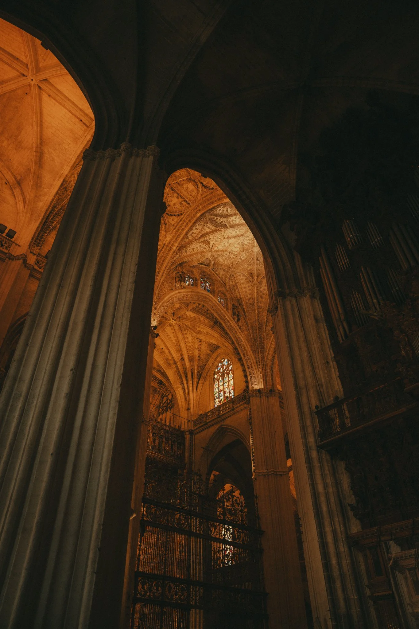 Interior view of a cathedral or church with tall columns, ornate ceilings, stained glass windows, and warm lighting.