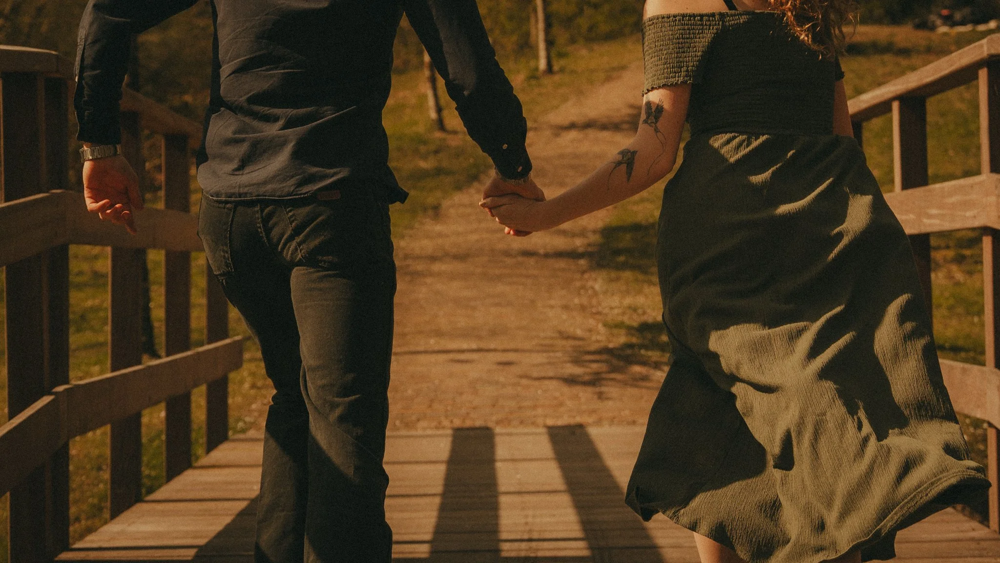 A man and woman holding hands on a wooden bridge during sunset, with the woman wearing a black dress and the man in dark clothing.