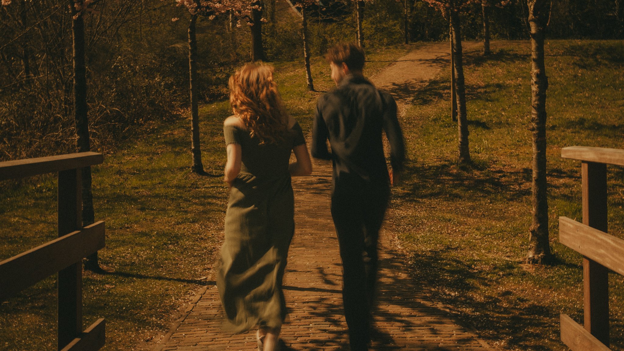 A man and woman walking together on a narrow wooden pathway surrounded by trees, at night, illuminated by warm lighting.