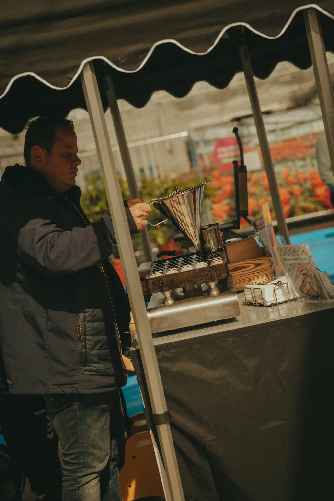 A man wearing a gray and black jacket operating a coffee or beverage stand with a metal funnel and a scale, under a black canopy.
