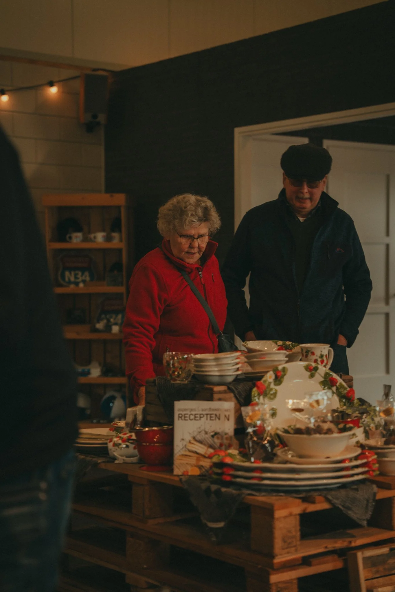 Two women, one elderly with gray hair and glasses, and the other younger wearing a black hat and sunglasses, are standing at a table with Christmas-themed dishware, serving or preparing food in a cozy, warmly lit room decorated with string lights.