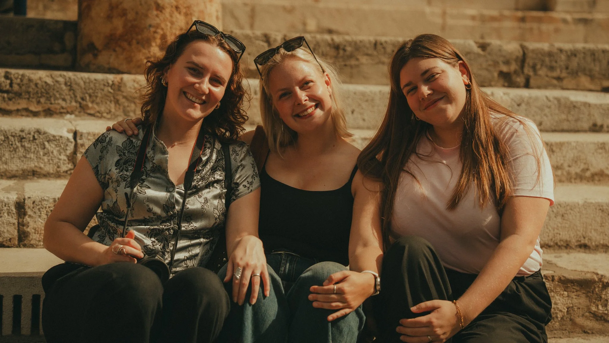 Three women sitting on stone steps, smiling, with glasses resting on their heads, during the evening.