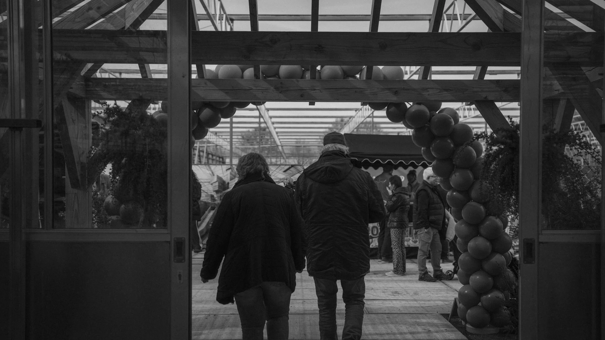 People entering a greenhouse or botanical garden through a glass door framed with a balloon arch, with other visitors inside.