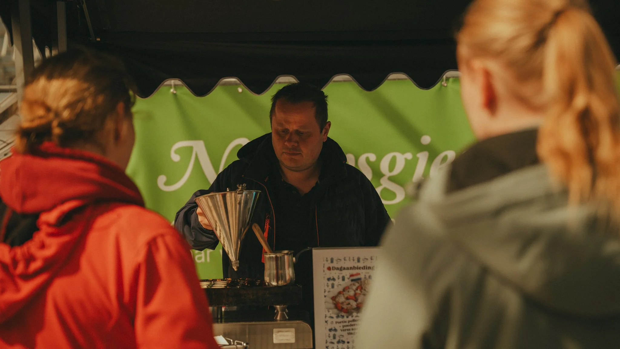 A man is serving food at a food stand with a green sign behind him, while two women stand in front of the stand, one in a red jacket and the other in a gray hoodie.