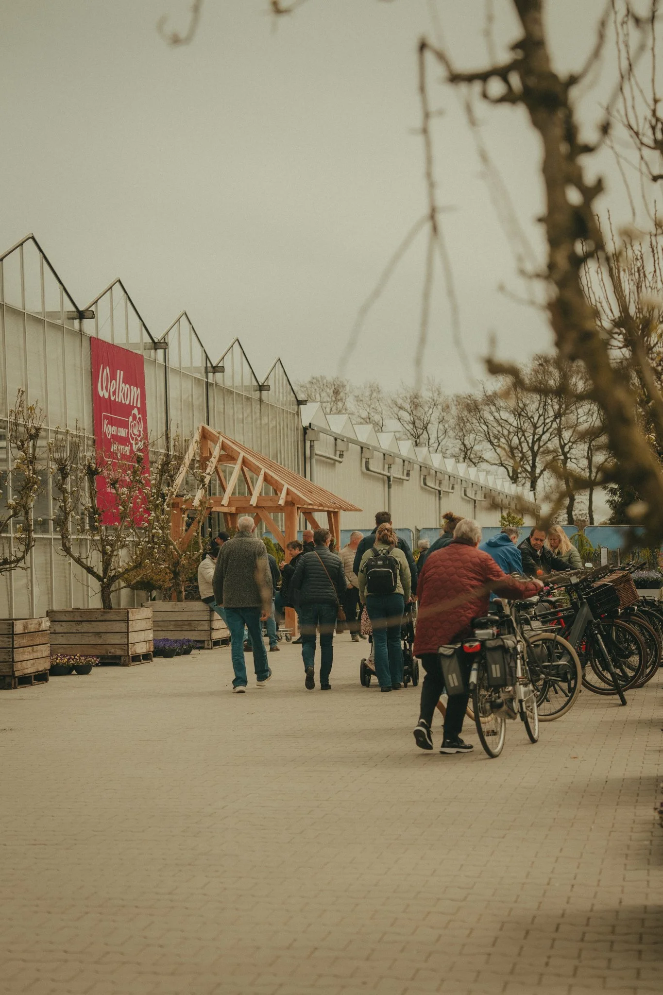 People gather outside a greenhouse with a red sign that says "Welkom" in Dutch. There are bicycles parked and some trees in the background, with a few people walking and talking.