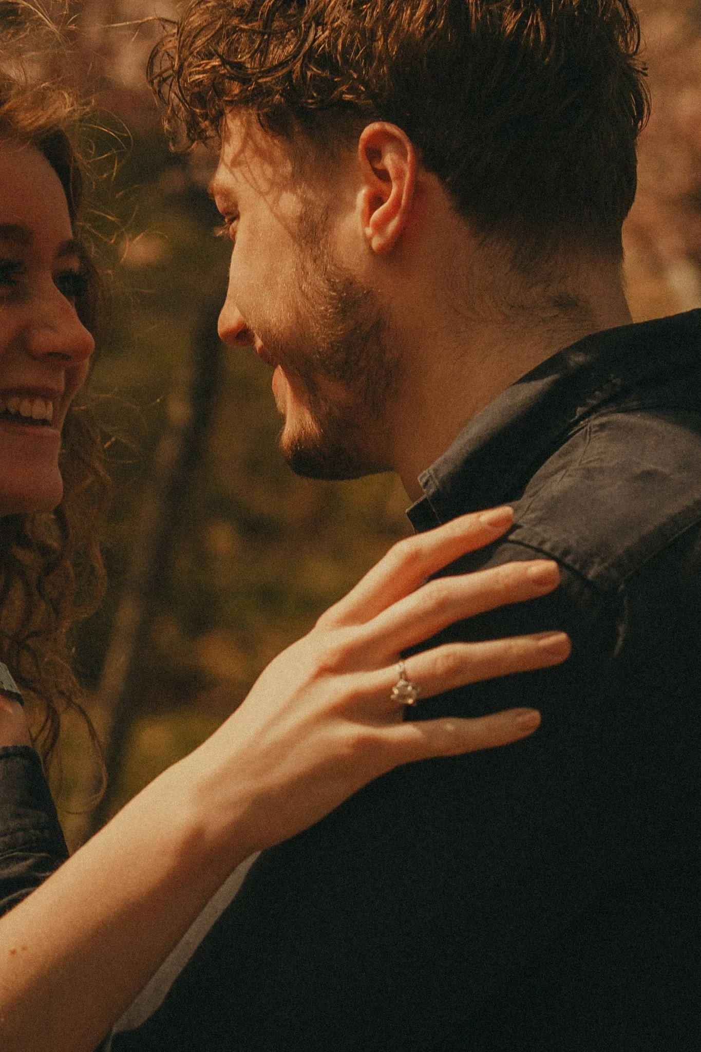A close-up of a couple smiling and looking at each other, with the woman's hand resting on the man's shoulder, both appearing happy and affectionate in an outdoor setting.