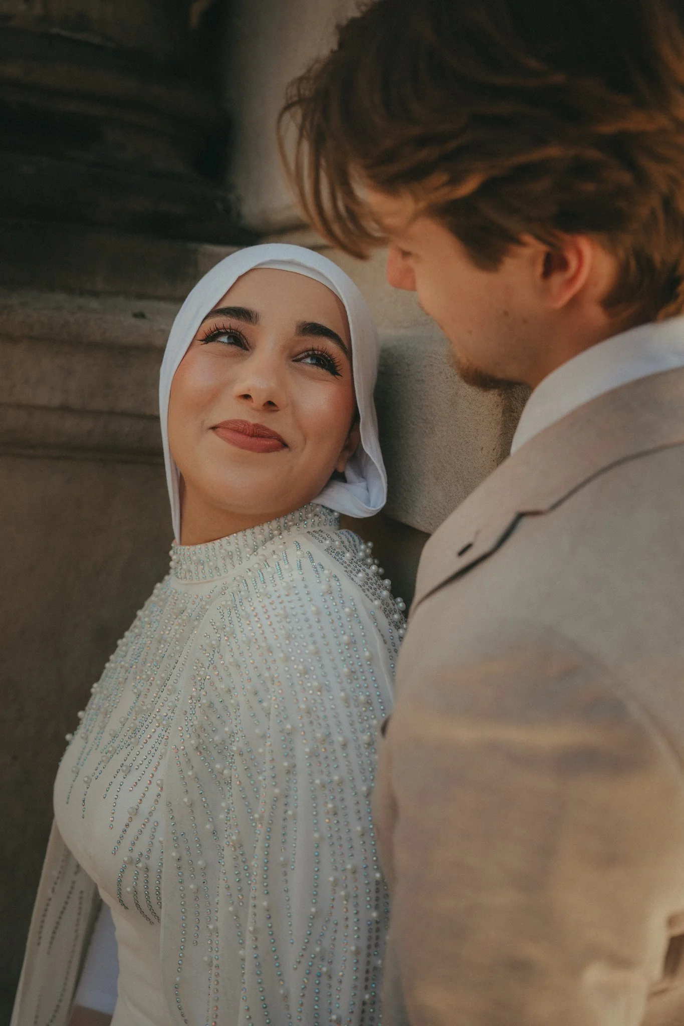 A woman wearing a hijab and a dress with decorative beads looking at a man, both smiling. They are in an outdoor setting with stones in the background.