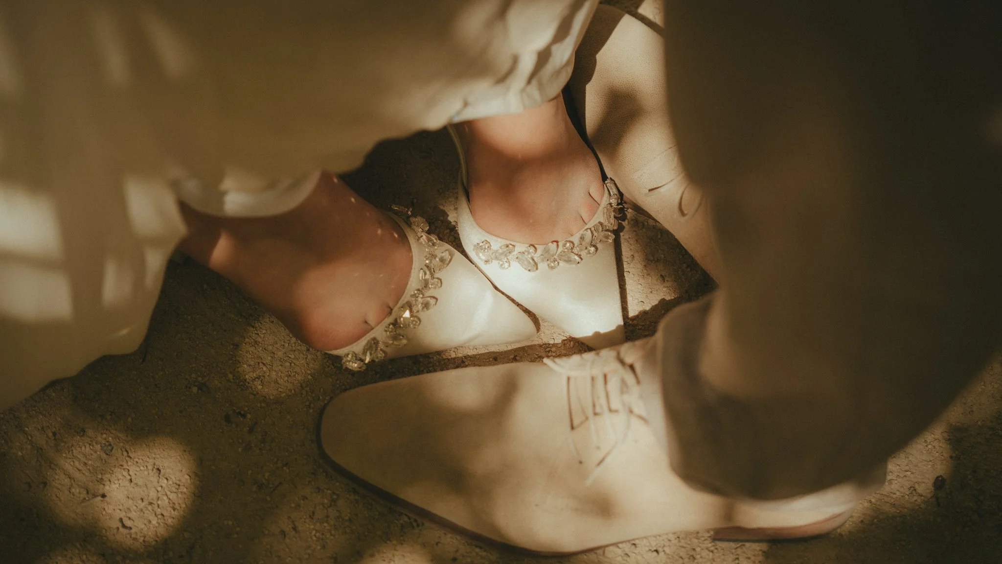 A person's feet with beige high heels with jeweled embellishments are shown in a narrow, sandy space, viewed from above.