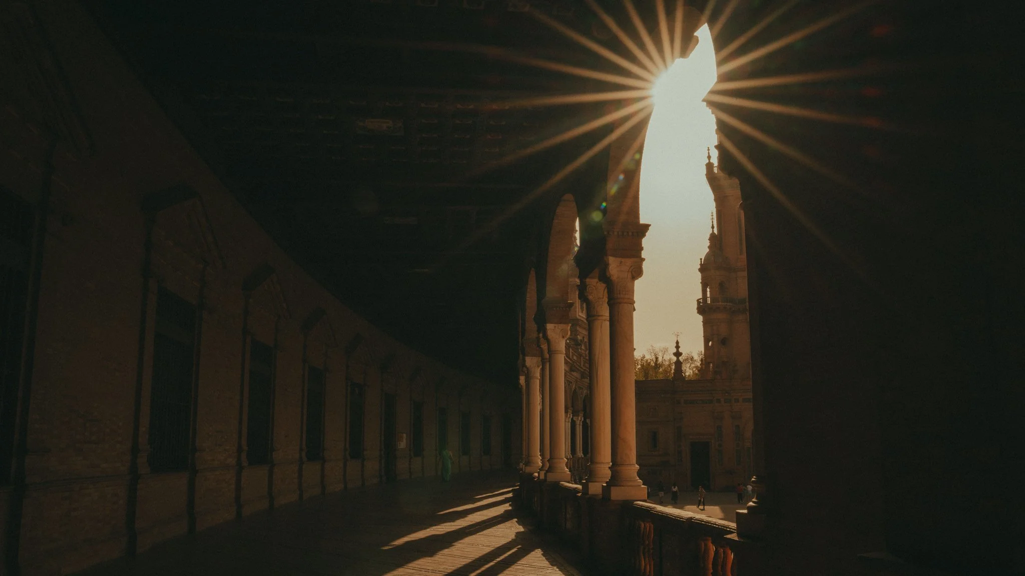 A view of an architectural structure with columns and arches, with sunlight shining through and creating rays; two small figures walk along a corridor.