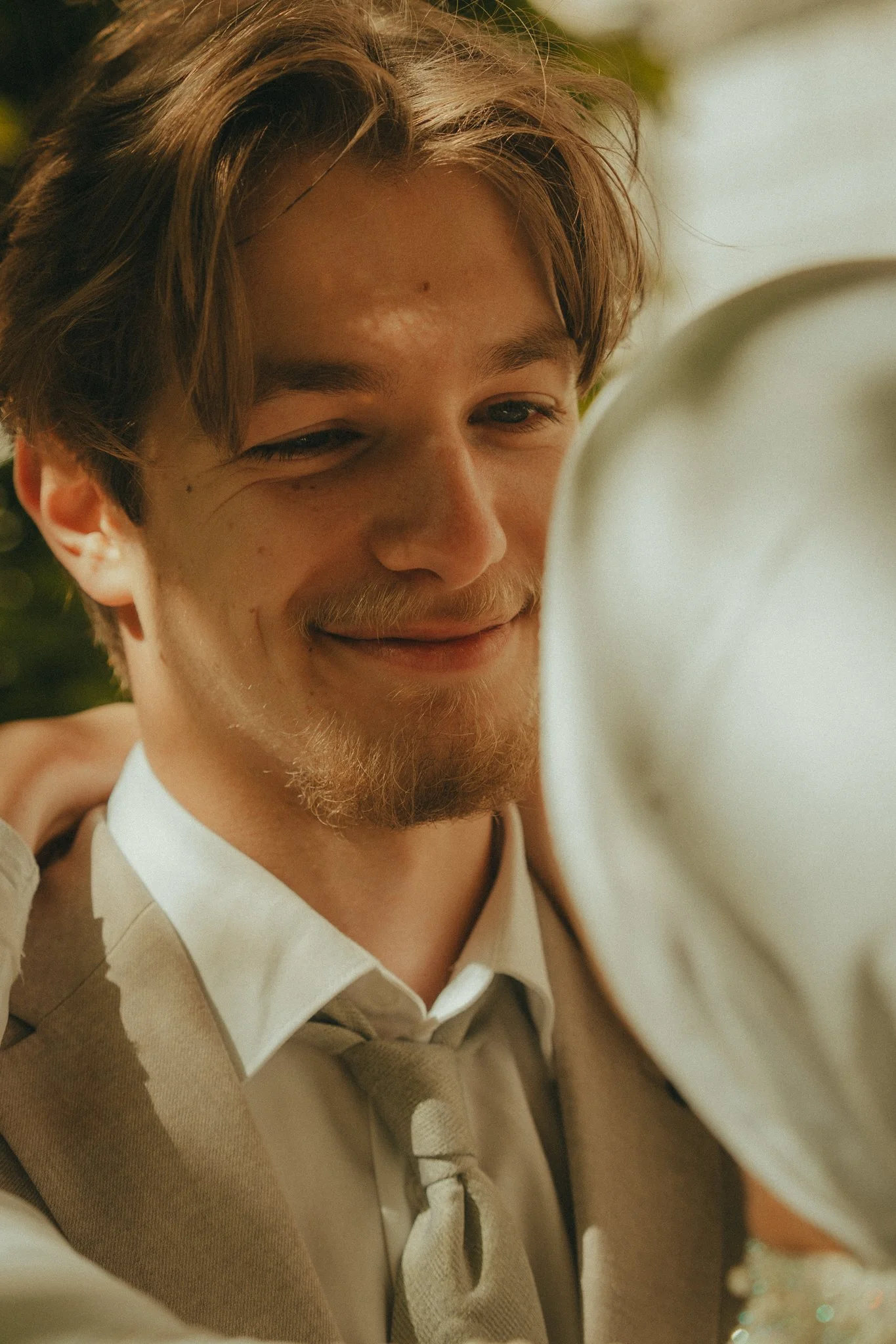 A young man with light skin, brown hair, and a beard, smiling while looking at a camera. He is wearing a white shirt and a beige sweater with a tie. The background appears to be outdoors with green foliage.
