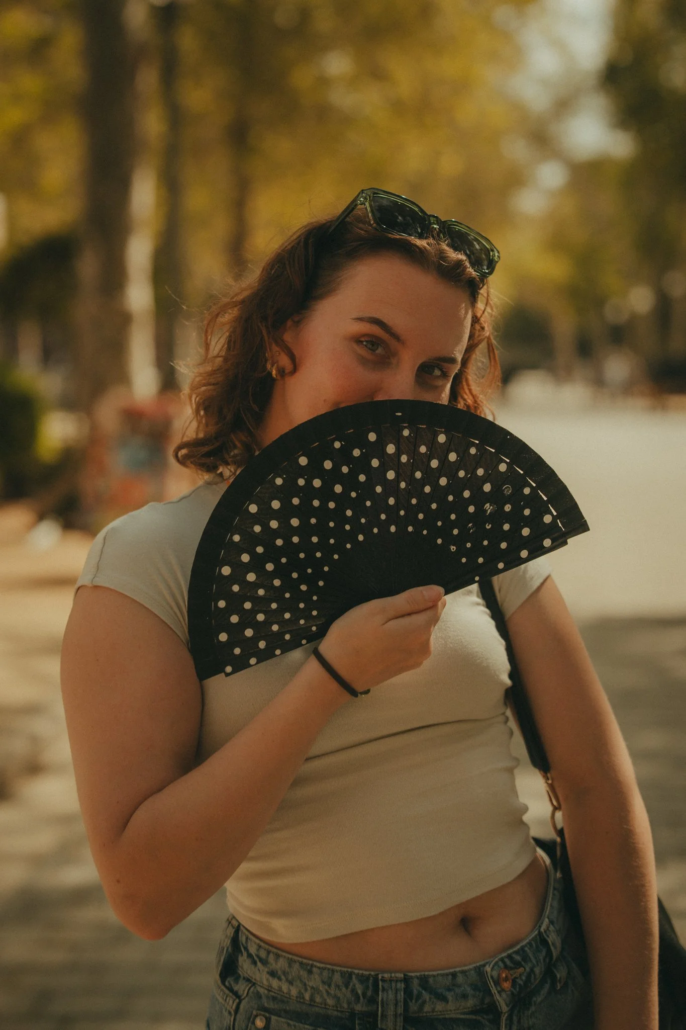 A young woman with curly brown hair wearing sunglasses on her head, a white crop top, and high-waisted jeans, stands outdoors holding a black fan with white polka dots partially covering her face, with trees and a pathway in the blurry background.