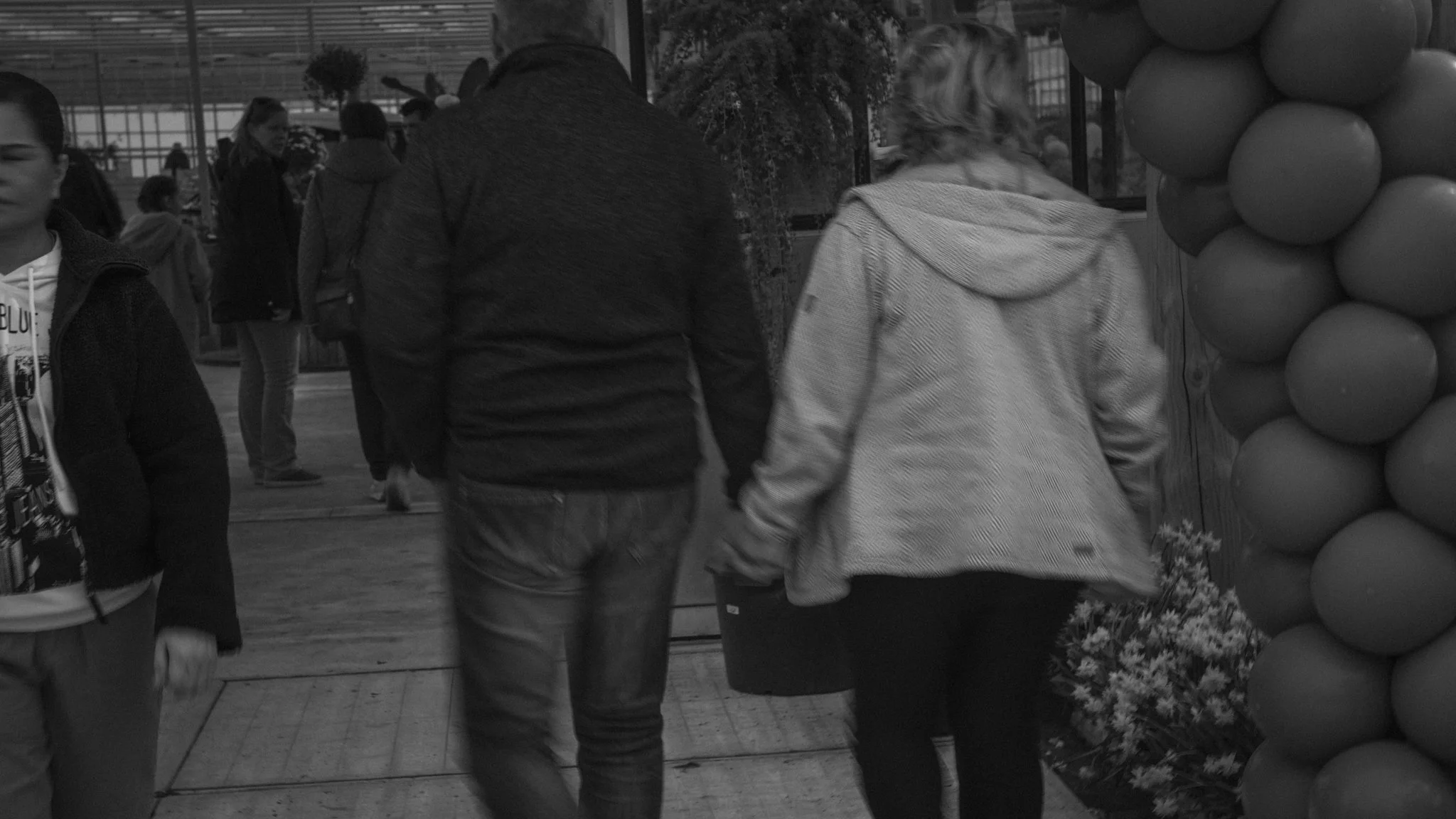A black and white photo of a group of people walking in an indoor space, possibly a mall or airport. In the foreground, a man and a woman are holding hands. The woman is wearing a patterned jacket, and the man is in a dark jacket. To the left, a youn