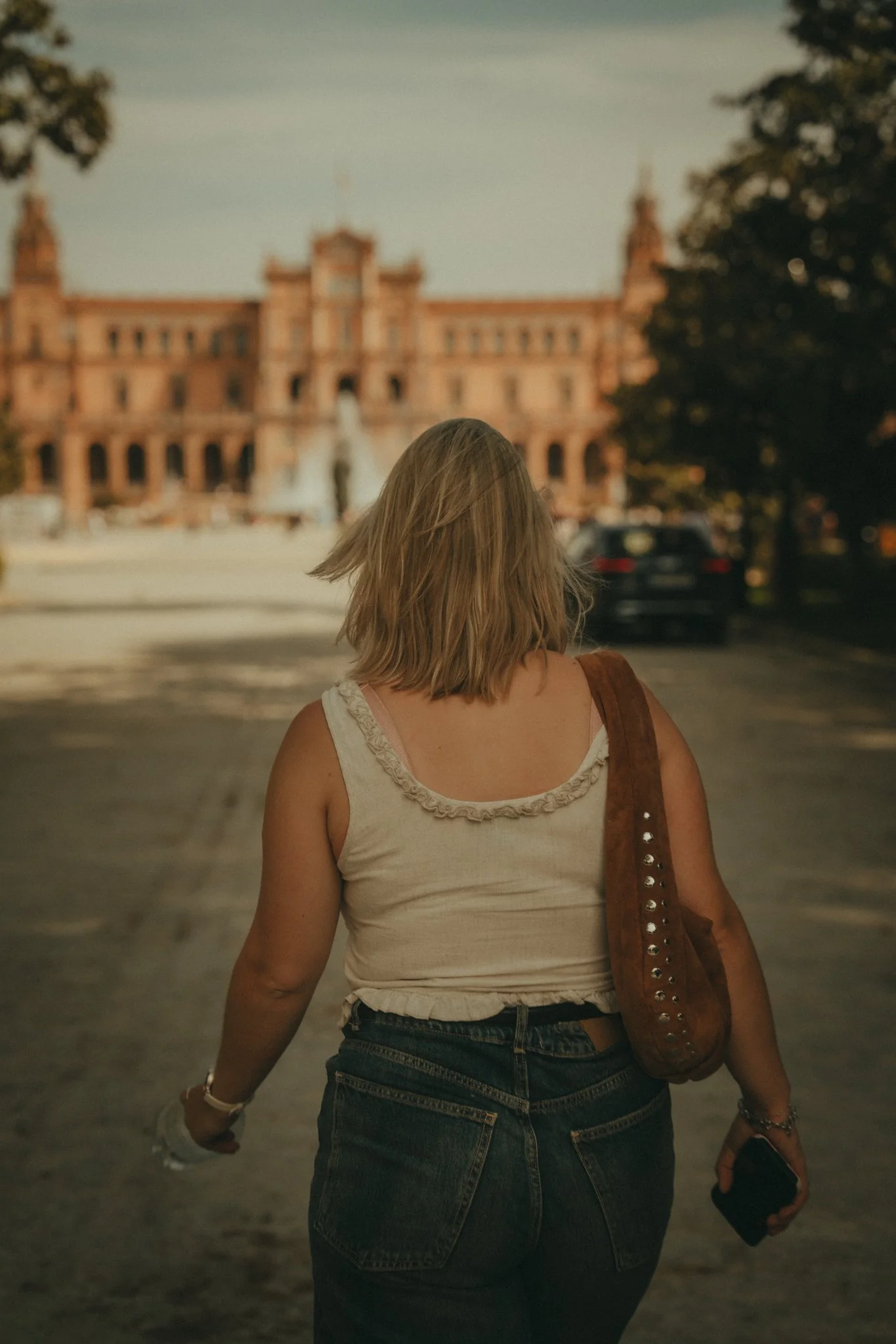 A woman with blonde hair, wearing a white tank top and jeans, walking on a street towards a large historic building in the distance, with trees on both sides.