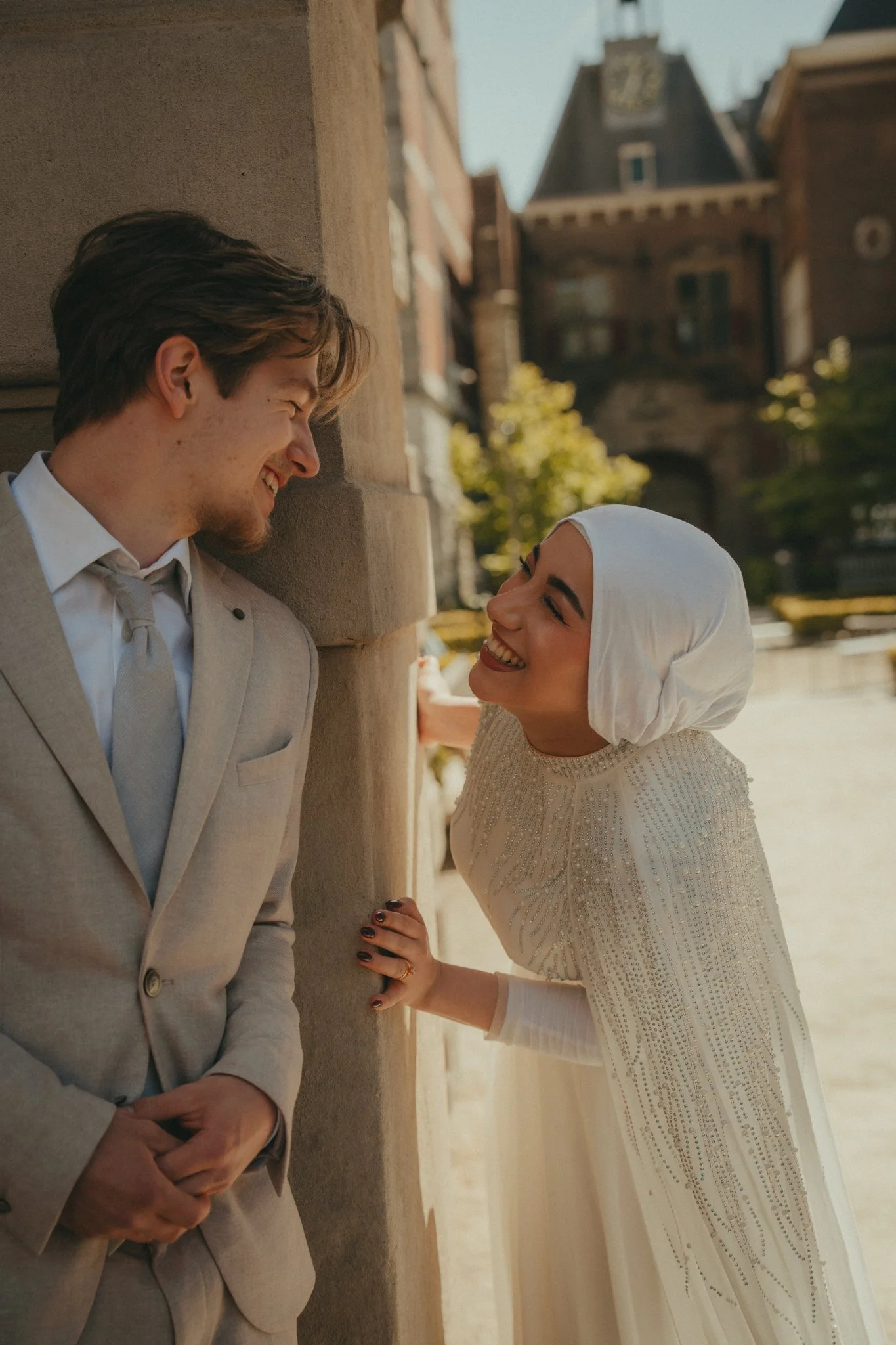 A man and woman sharing a lighthearted moment outdoors, leaning against a building, with a historic building and trees in the background.
