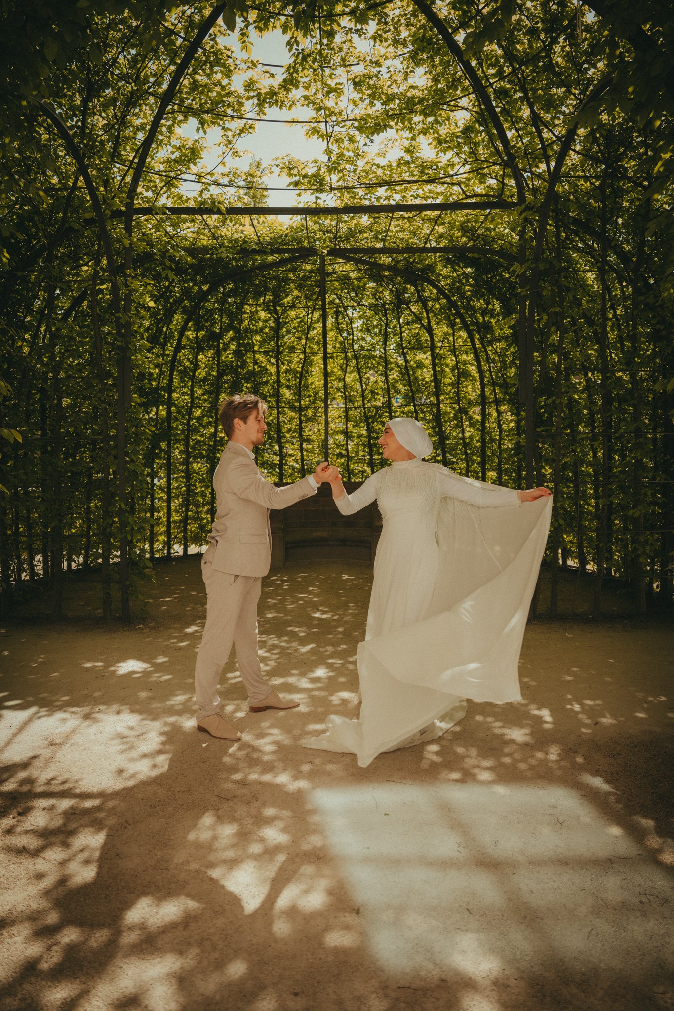A couple dancing together outdoors under a canopy of green leaves, holding hands and smiling at each other, with dappled sunlight casting shadows on the ground.