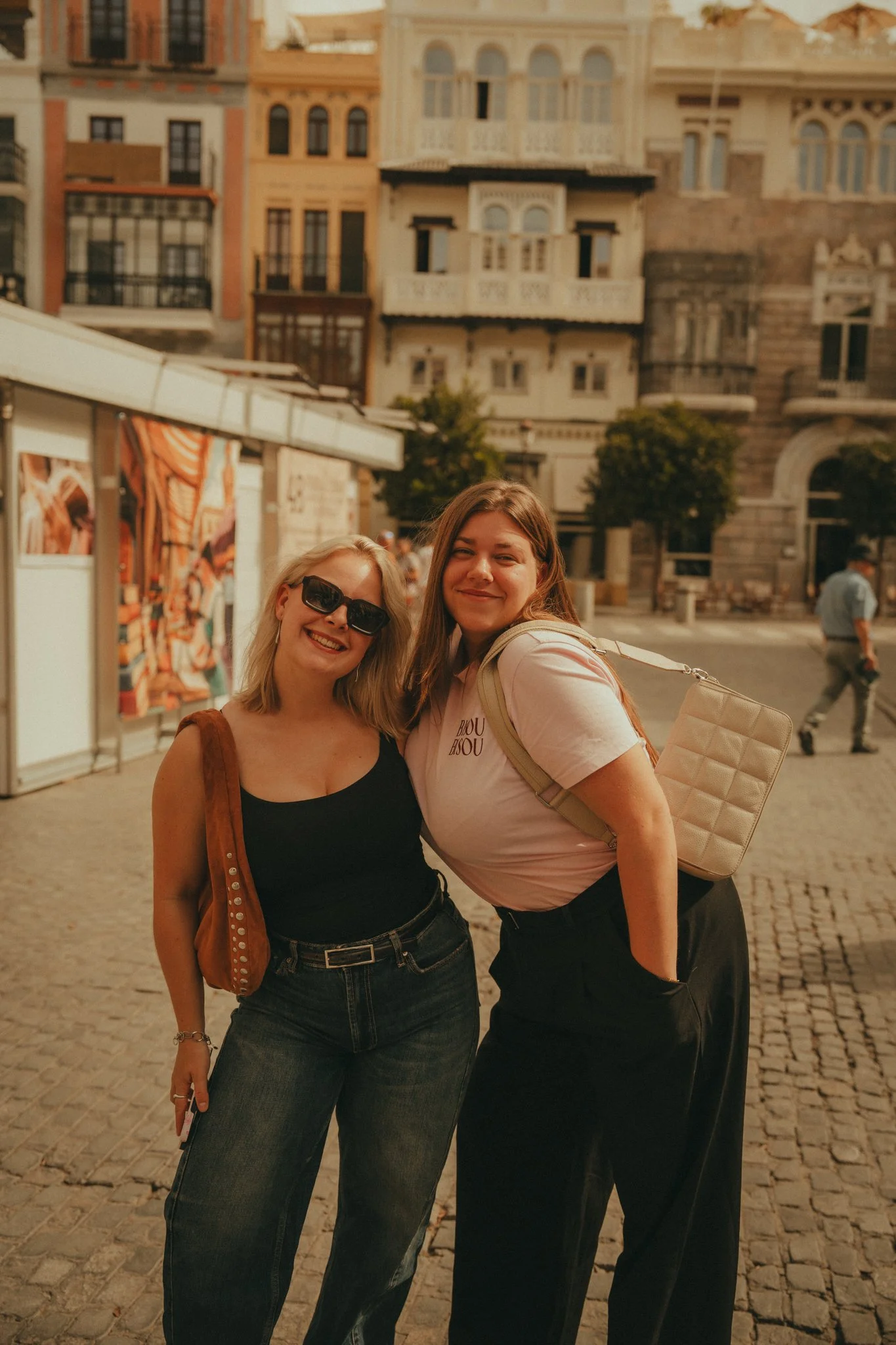 Two women smiling and standing close together on a cobblestone street with historic buildings in the background.