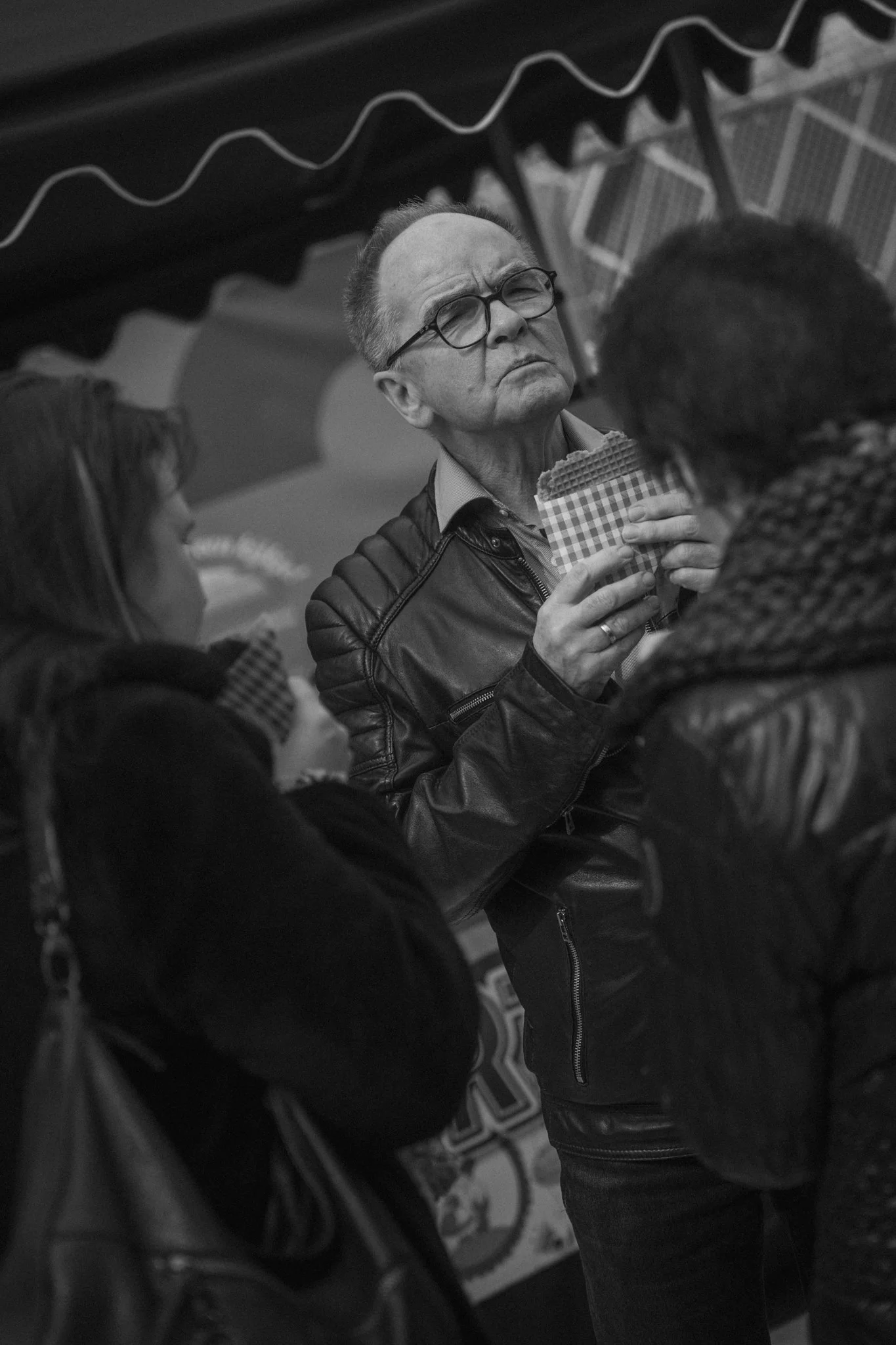 A man wearing glasses and a leather jacket is holding a waffle cone of ice cream with checkered paper wrapping, looking upwards, surrounded by two women in what appears to be a social setting.
