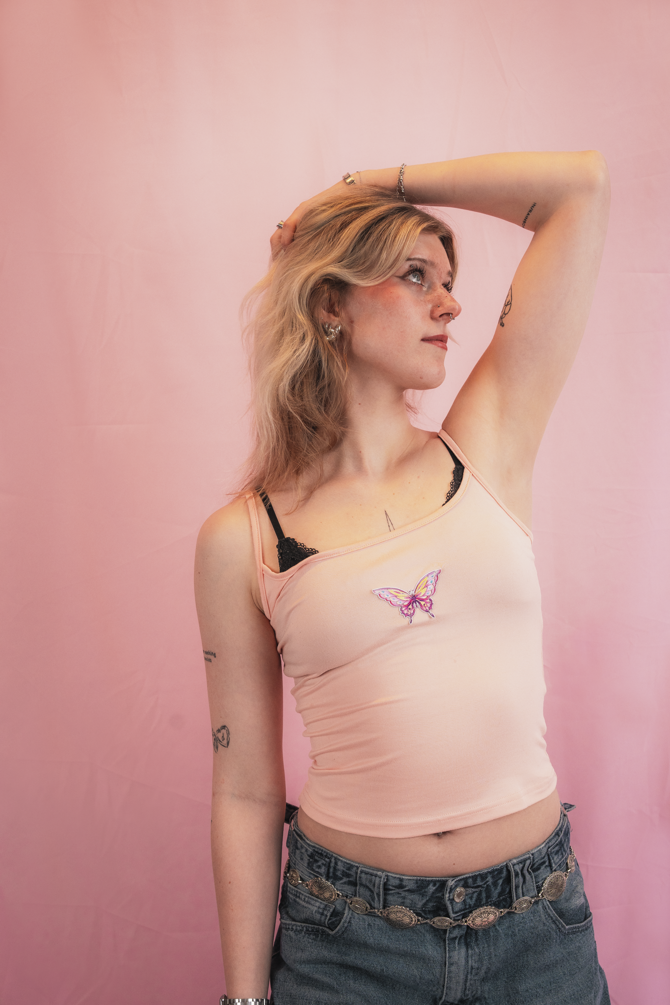 A young woman with blonde hair and tattoos on her arm, wearing a light pink tank top with a butterfly design, standing against a pink backdrop, looking upward with one hand on her head.